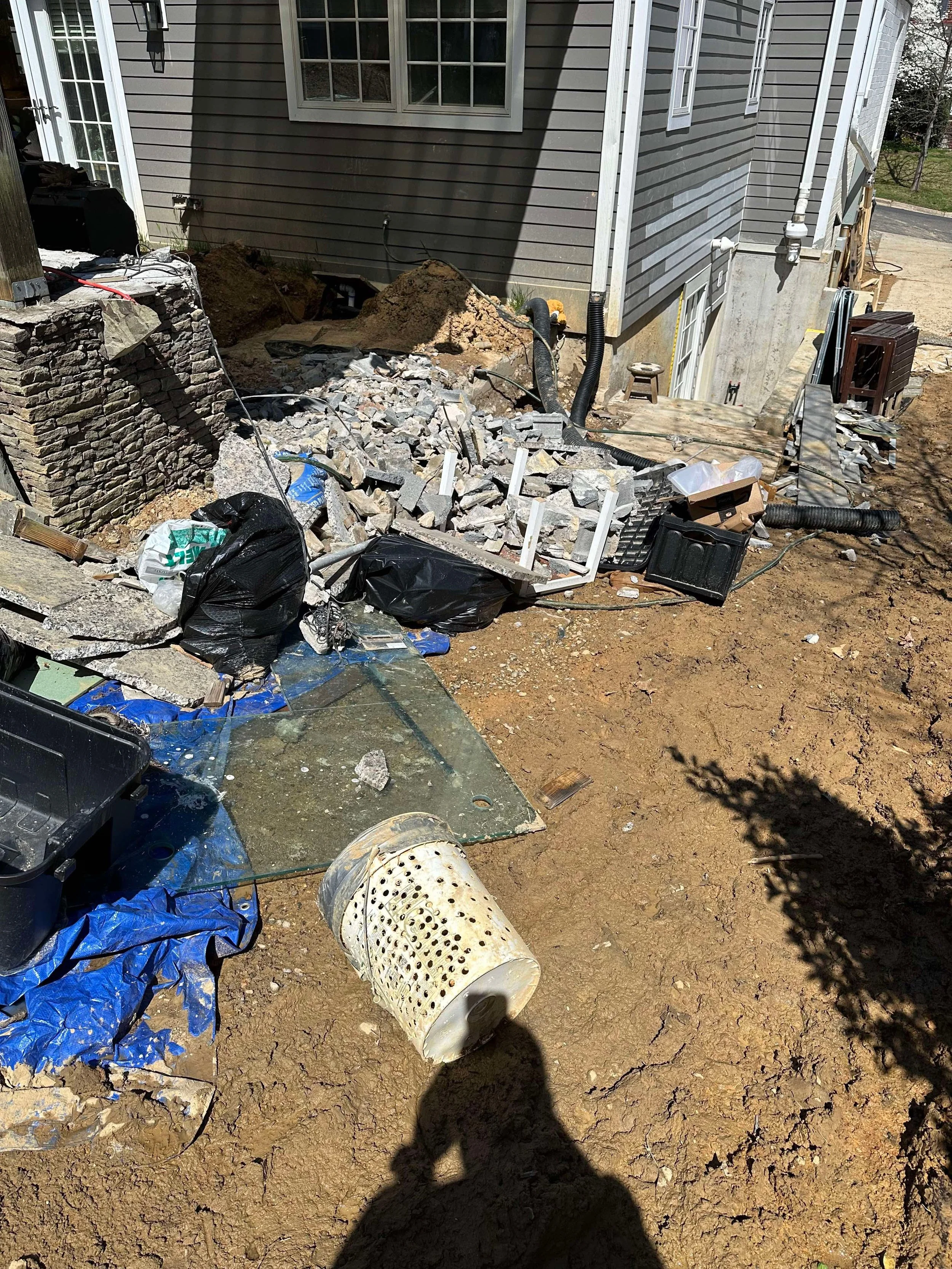 Construction site with scattered bricks, debris, black trash bags, a bucket, plastic sheets, and construction tools near a house with gray siding and a small set of stairs.