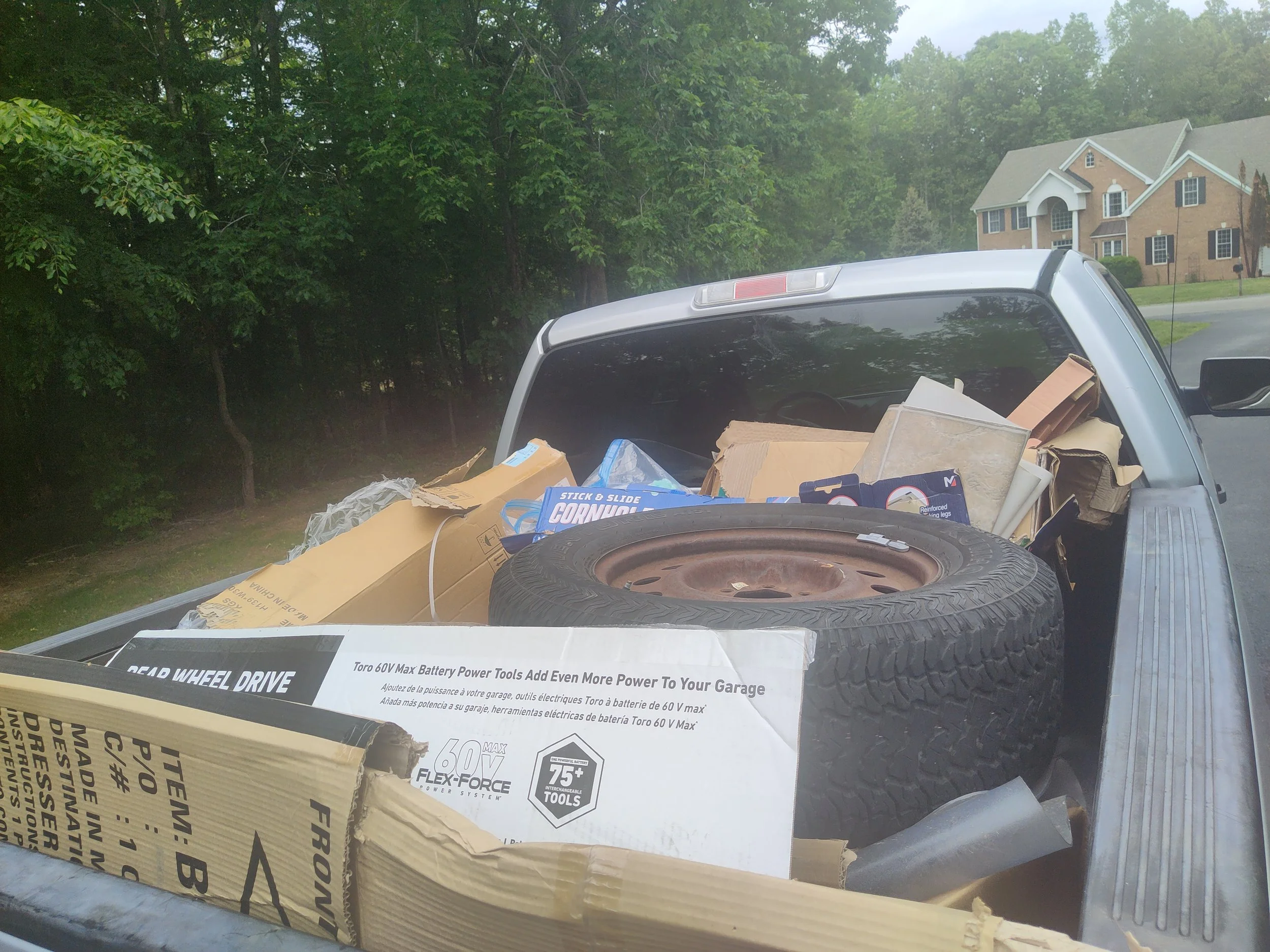 A truck bed filled with boxes, tools, and a spare tire, parked in a residential neighborhood with green trees and houses in the background.
