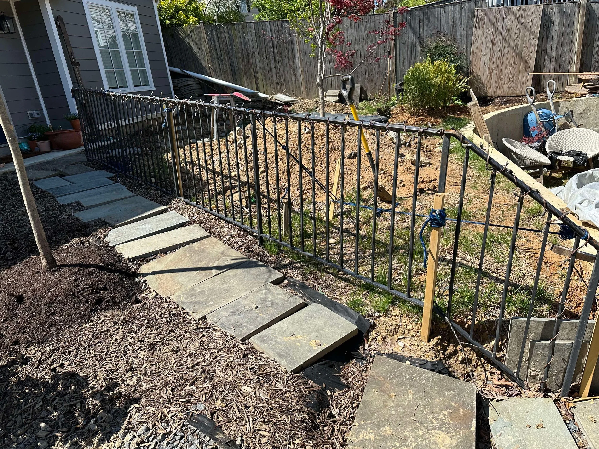 A backyard under construction with a stone pathway, a newly installed metal fence, and a garden area with chairs and tools.