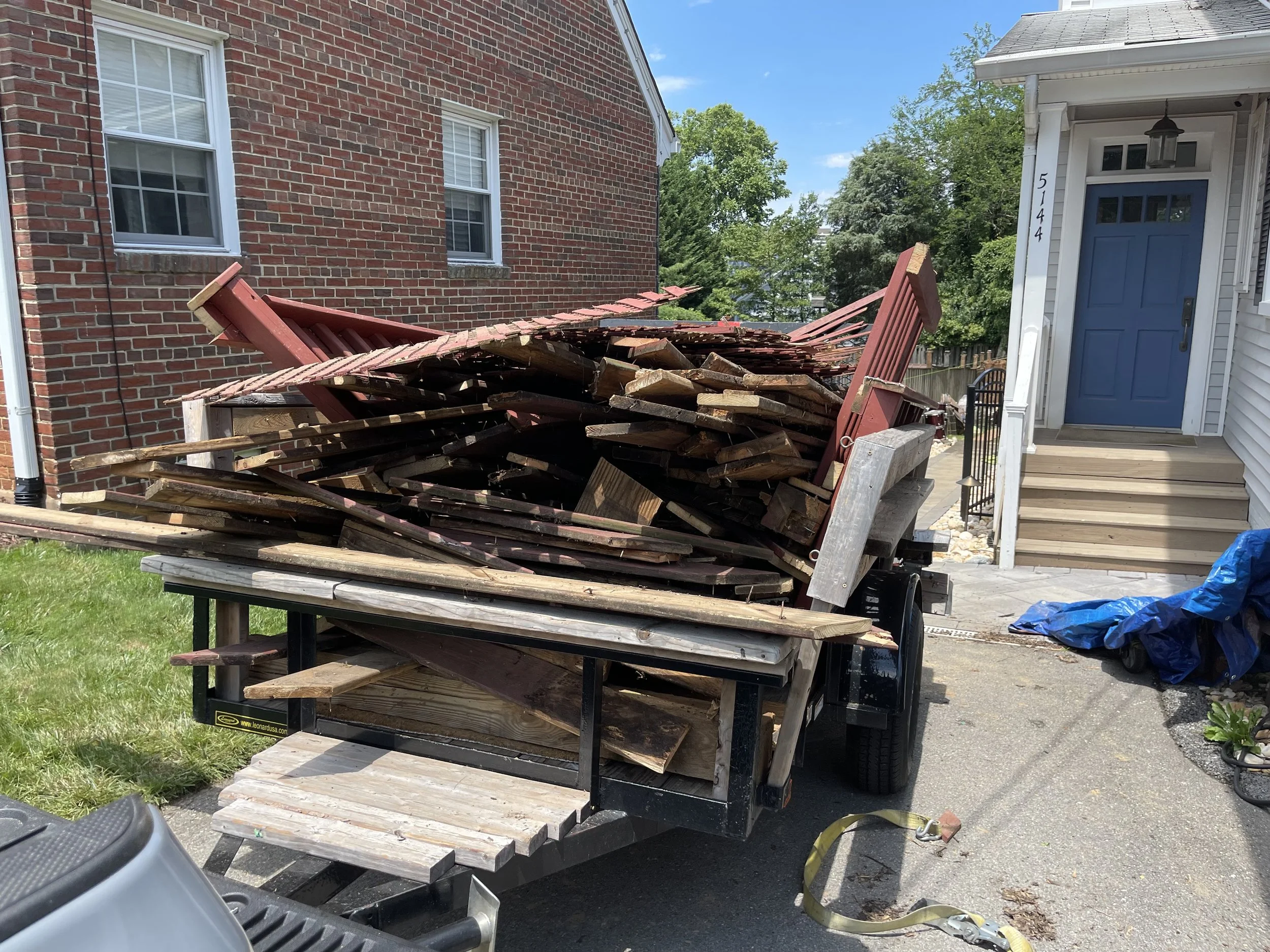 Trailer filled with wooden planks and debris parked in front of a residential house with a blue front door and stairs.