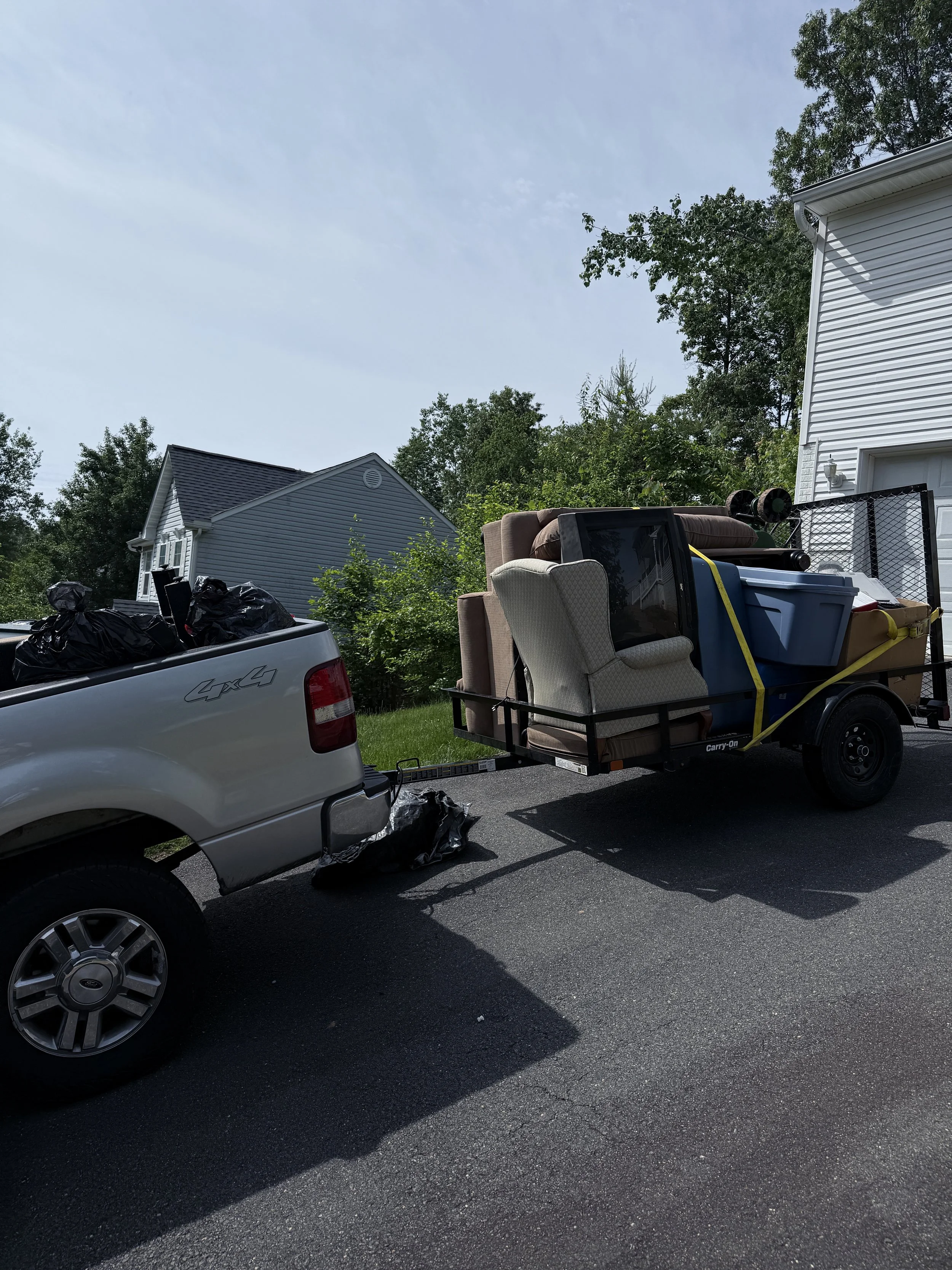 A pickup truck towing a trailer loaded with furniture and household items, parked on a residential driveway with houses and trees in the background.