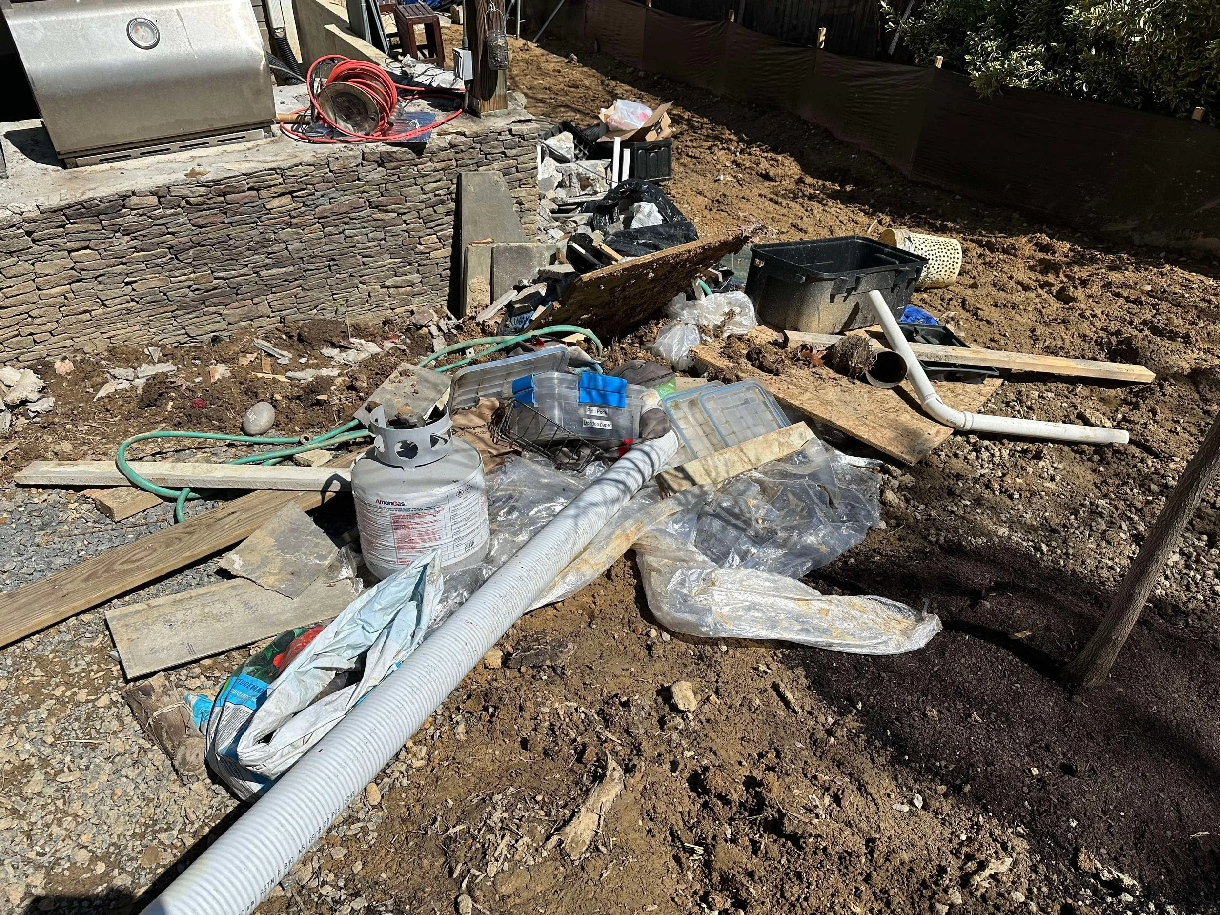 Construction site with scattered debris including pipes, plastic containers, tools, and building materials on dirt ground in front of a stone wall.