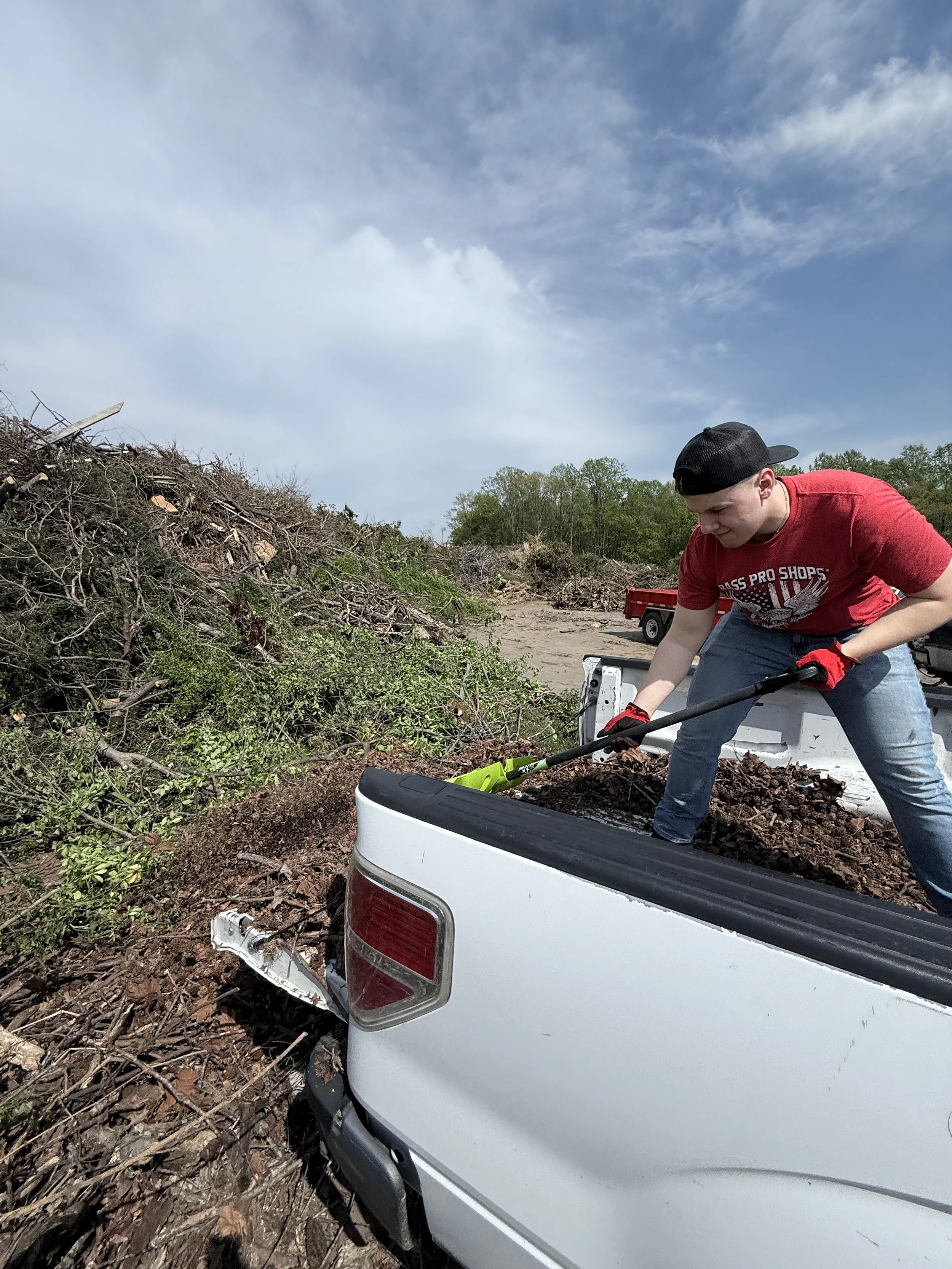 A man in a red T-shirt and a black cap using a rake to clear debris from the bed of a white pickup truck at a landscape with cleared land and scattered bushes under a blue sky.