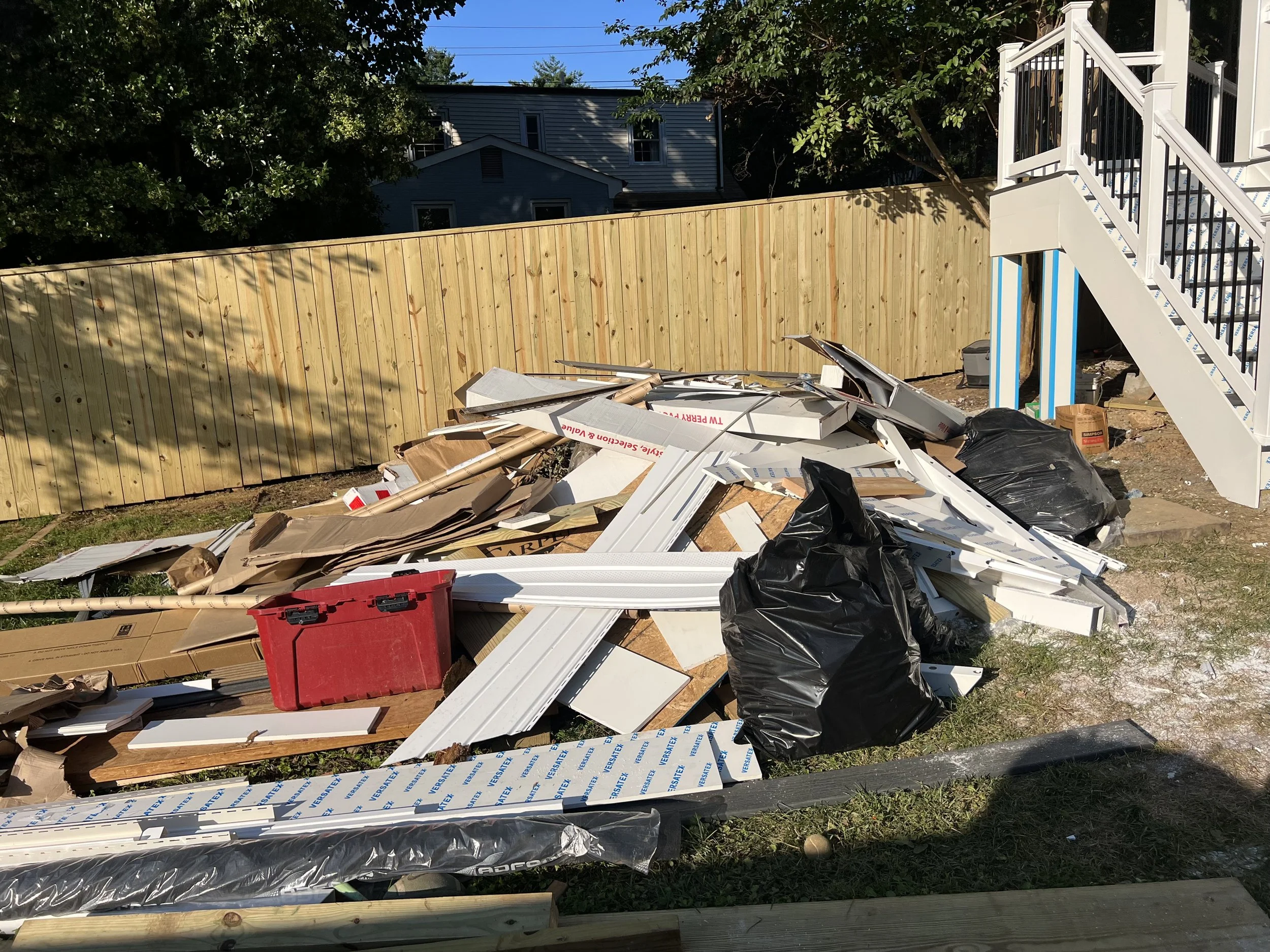Construction debris including cardboard, black trash bags, white metal parts, and tools scattered on the ground outside a house with a wooden fence and a staircase.