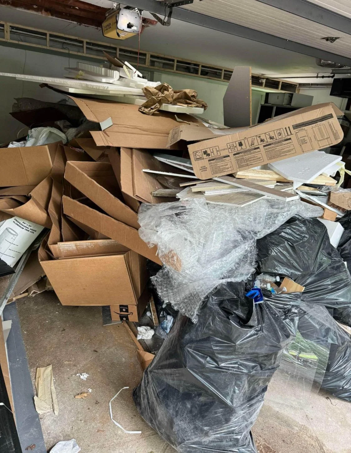 A cluttered pile of cardboard boxes, black trash bags, and packaging materials in a garage or storage area.