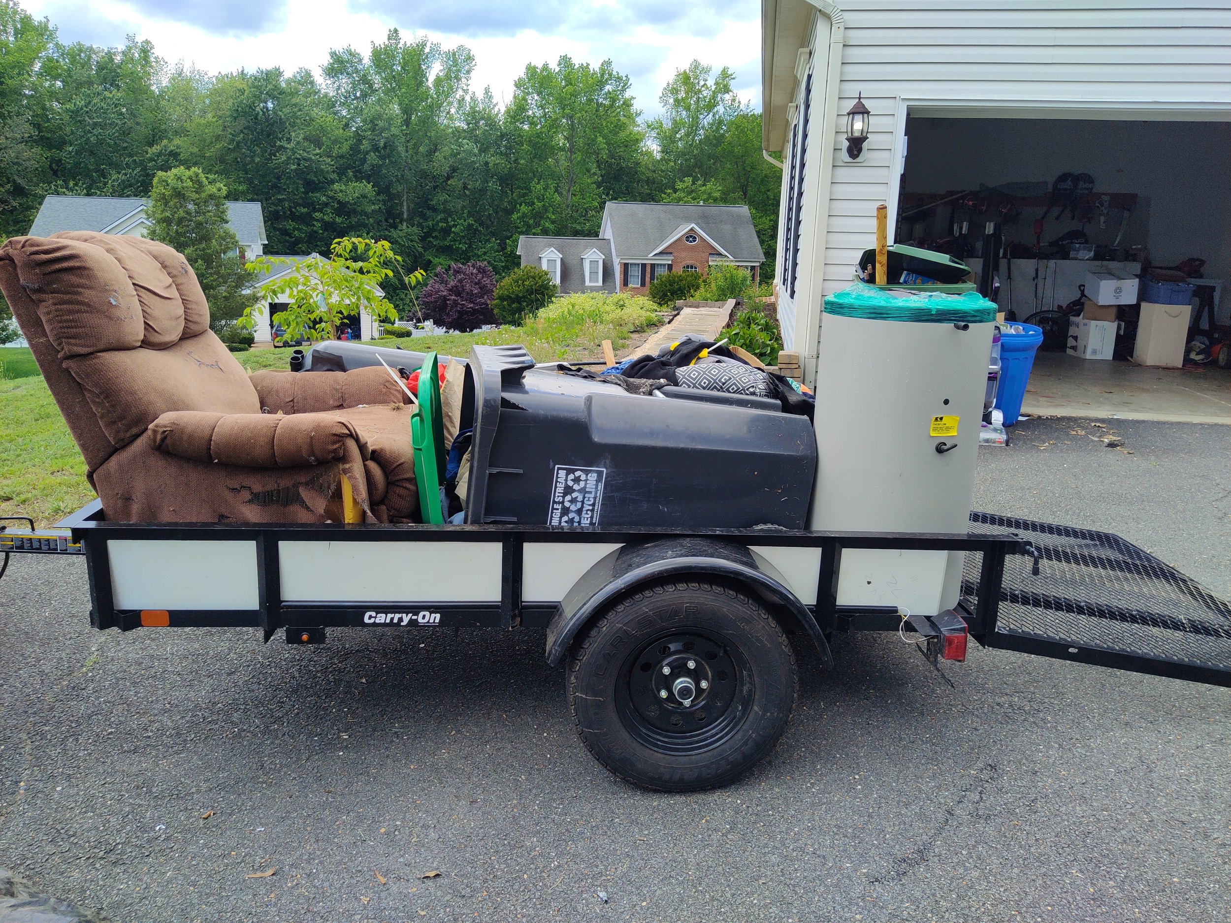 A small trailer parked in a driveway loaded with furniture and household items, including a brown recliner, a trash bin, and storage containers, with a garage and suburban houses in the background.