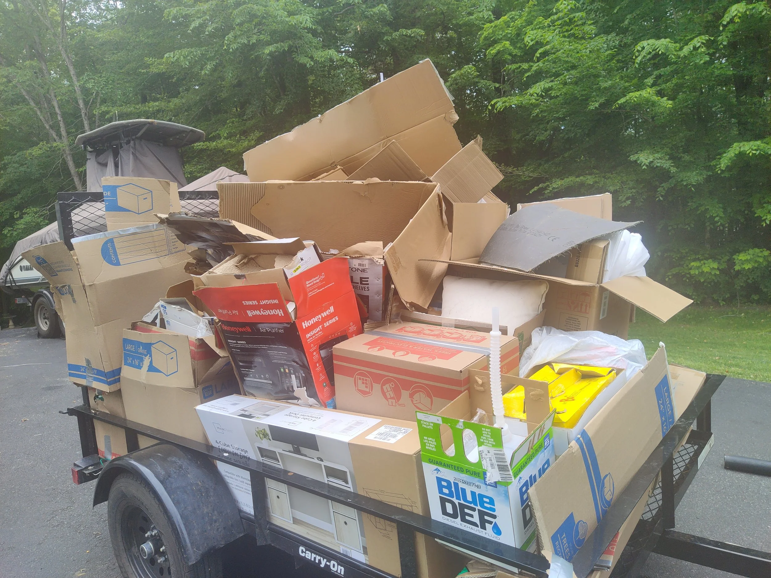 Trailer loaded with partially crushed cardboard boxes and packaging materials parked in a driveway surrounded by green trees.