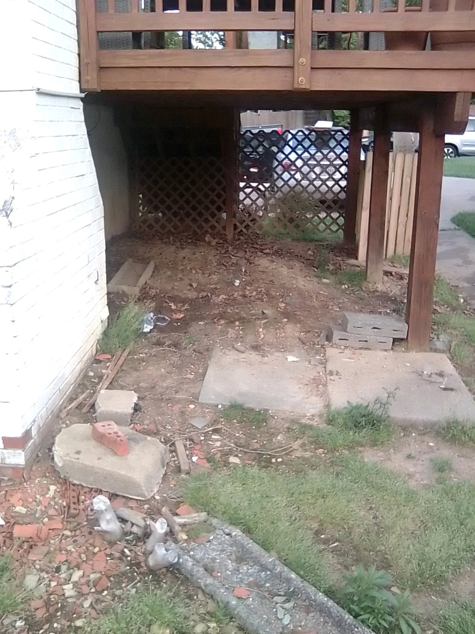 Rear view of a house with an elevated wooden deck, a small dirt area underneath, a lattice fence, and a stone pathway with some grass and plants.
