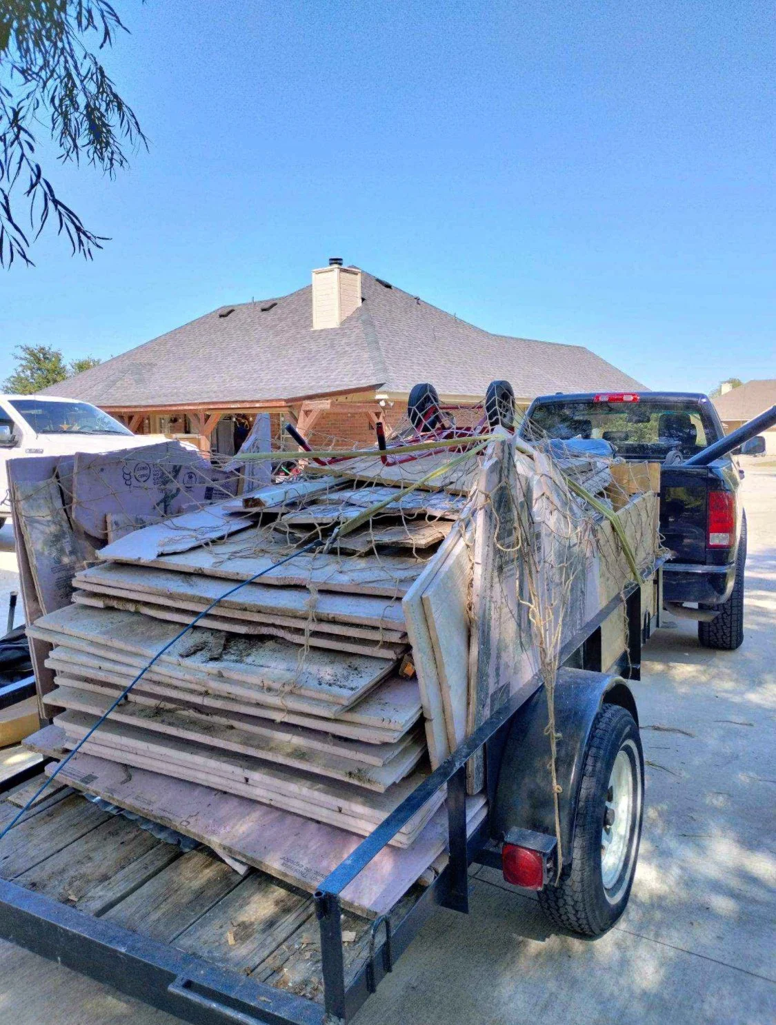 A trailer filled with construction debris, including plywood and drywall, attached to a black pickup truck in a driveway outside a house with a gray roof and brick chimney.