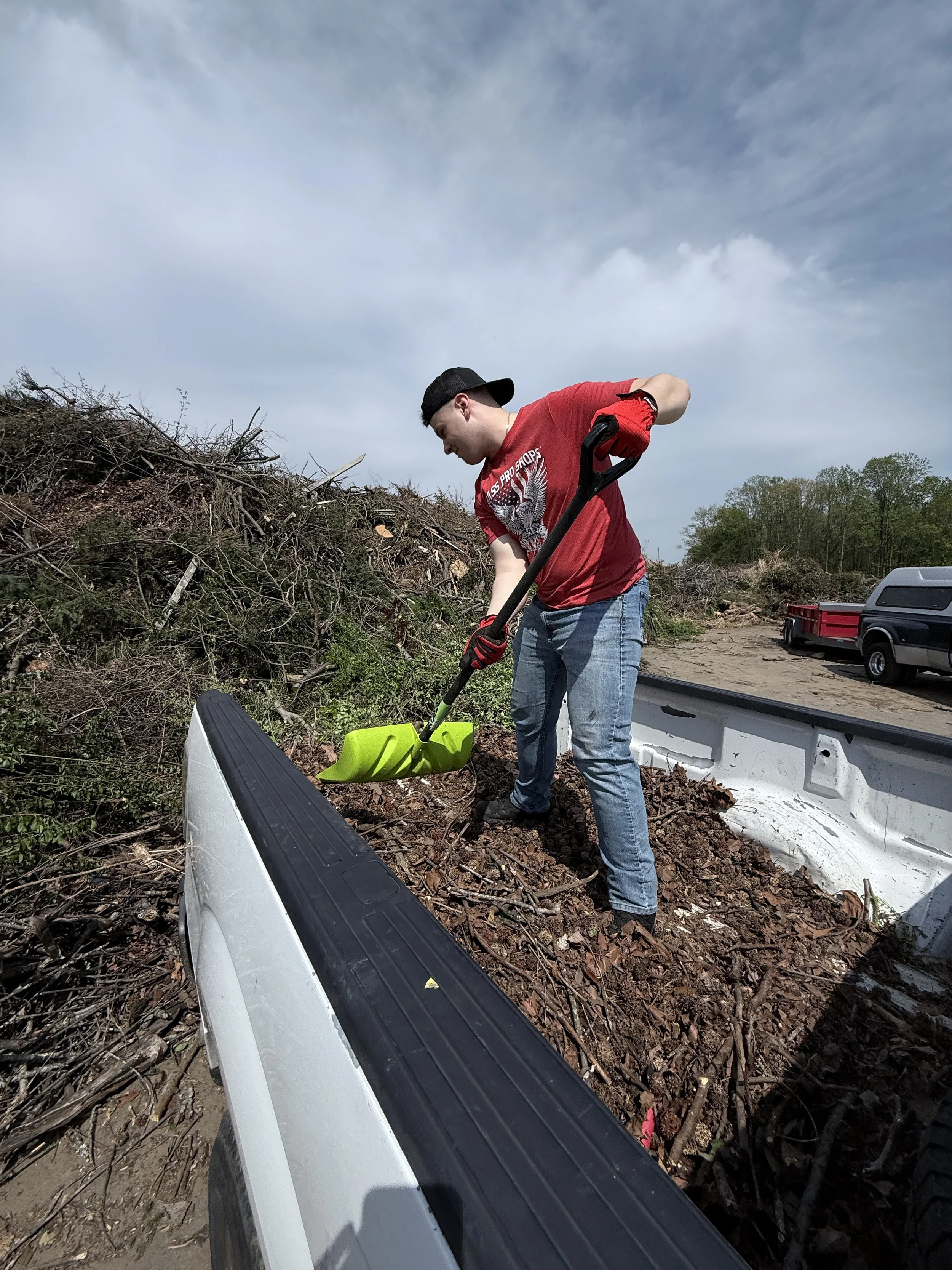 A young man wearing a red T-shirt, blue jeans, red gloves, and a black cap is shoveling dirt and debris into the bed of a pickup truck during outdoor cleanup.