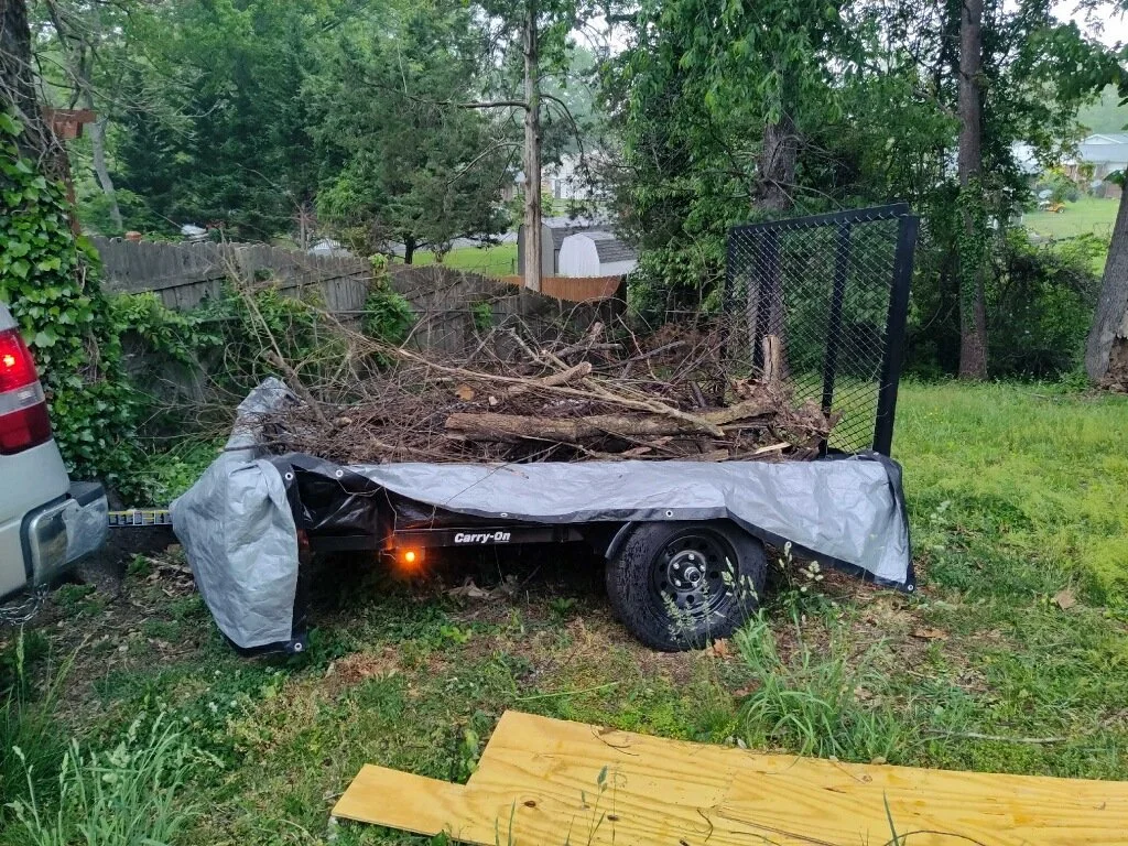 A trailer filled with tree branches and debris is attached to a vehicle in a backyard with green grass and trees.