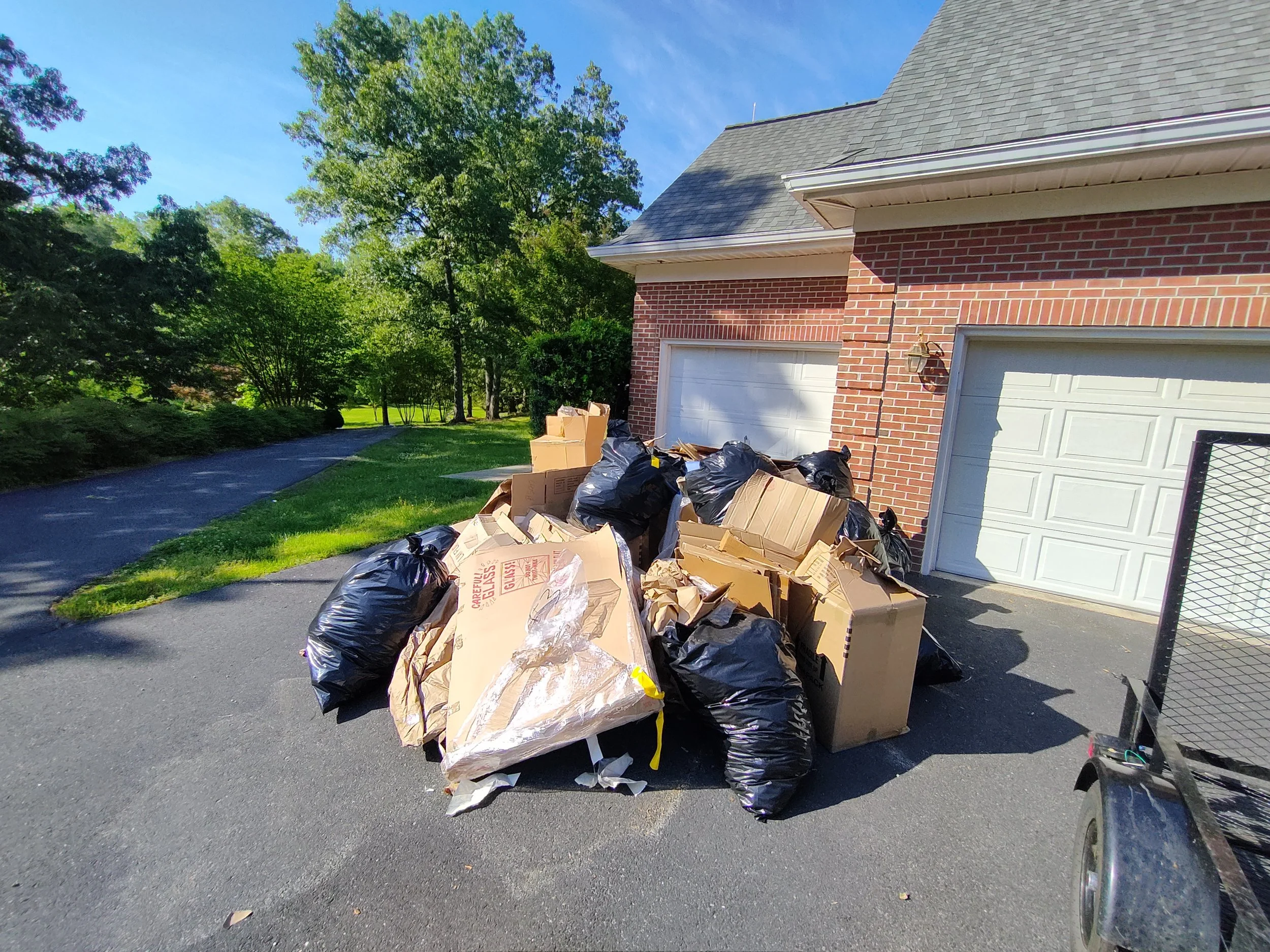 Overflowing trash bags and cardboard boxes in driveway near house with brick exterior and white garage doors, sunny day with green trees in background.
