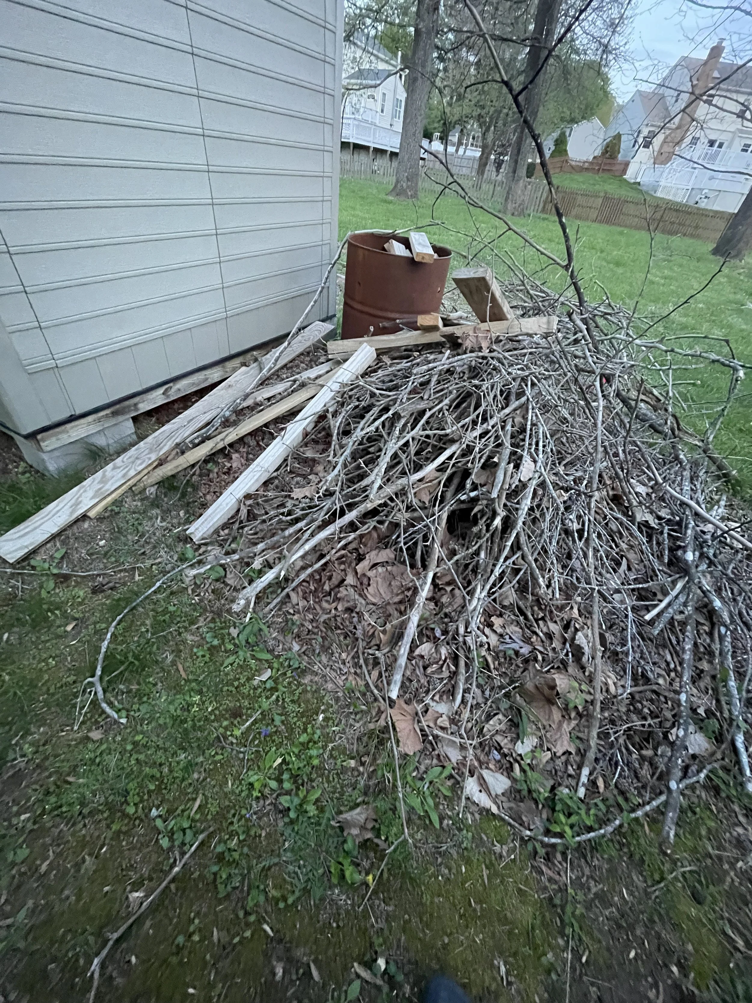A pile of dry branches, leaves, and small sticks next to the side of a house with horizontal siding, with some wooden planks and a rusty metal barrel on top of the pile. There are trees and other houses in the background.