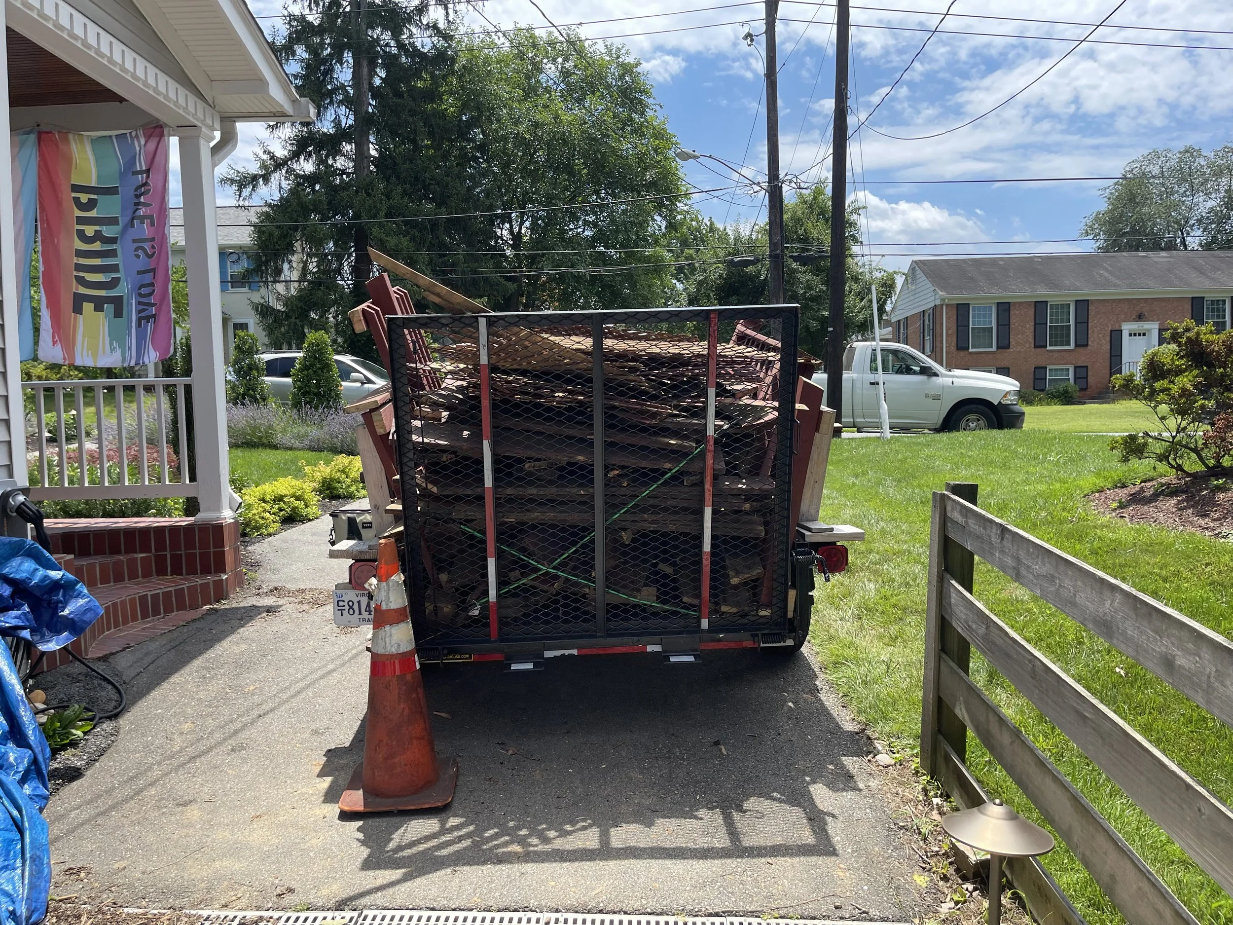 A trailer filled with wooden planks parked on a driveway next to a house, with an orange traffic cone in front.