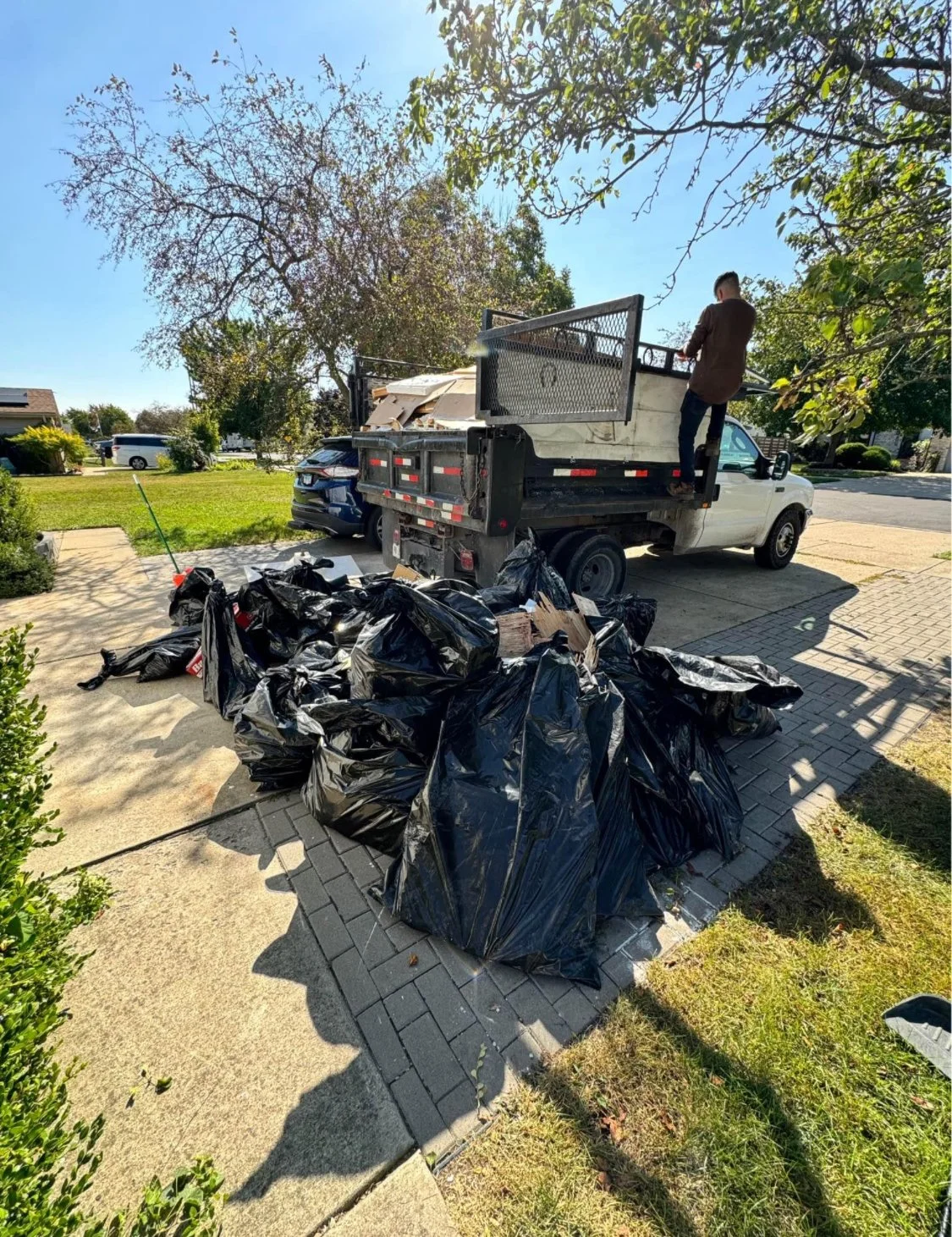 Person standing on the bed of a flatbed truck, unloading black trash bags filled with yard waste or trash, parked on a driveway in a suburban neighborhood.