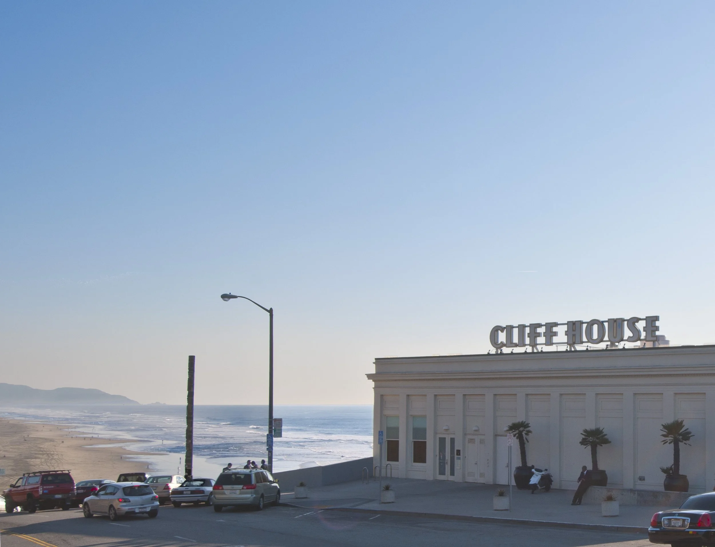 A coastal scene showing a parking lot with cars, street lamps, a building with a sign that reads 'CLIFFHOUSE', palm trees, and the beach and ocean in the background on a clear day.