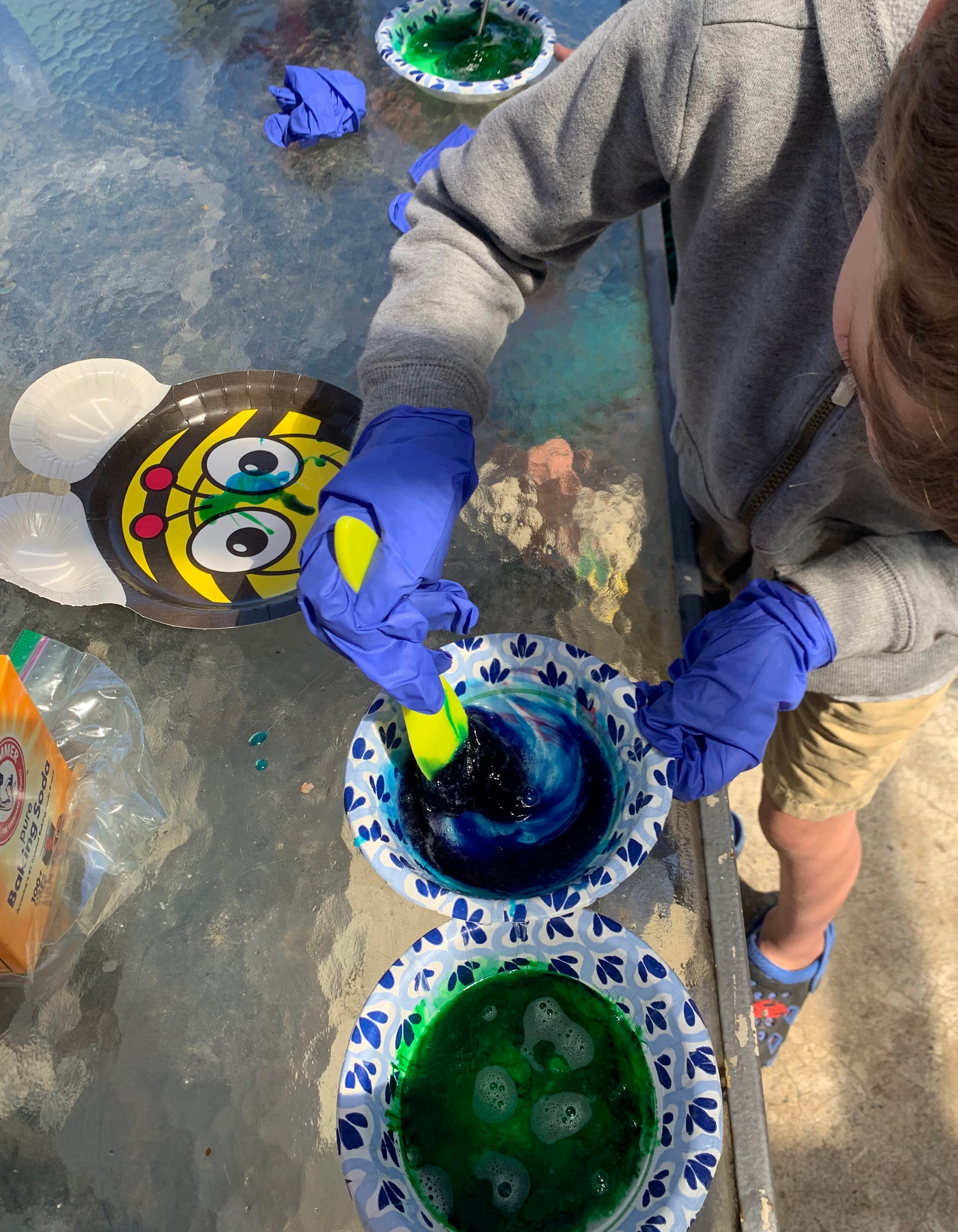 Children wearing gloves mixing green slime in paper bowls on a table.