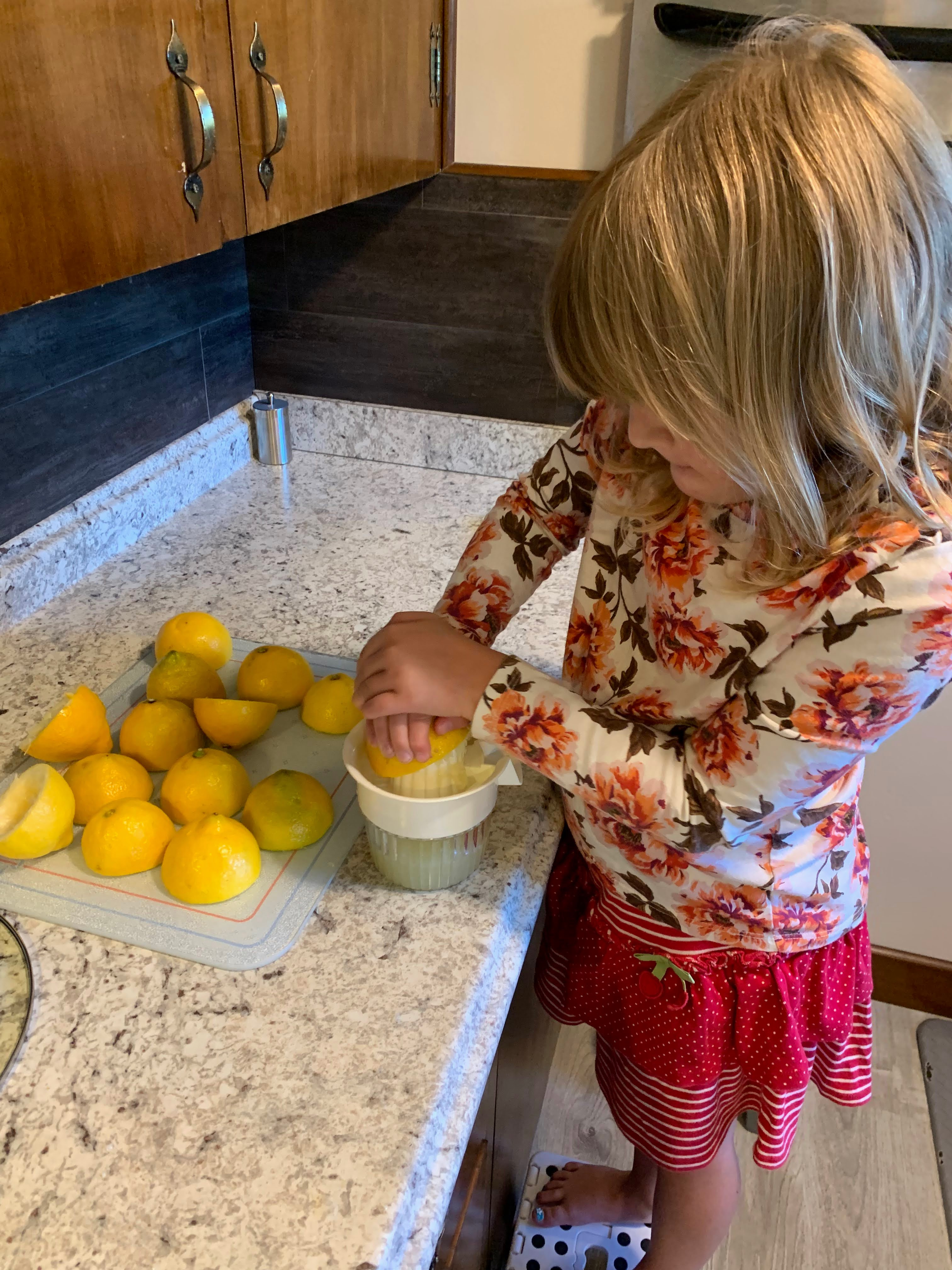 A child with blonde hair, wearing a floral long-sleeve shirt and red polka-dot skirt, is standing on a step-stool while juicing lemons for lemonade.