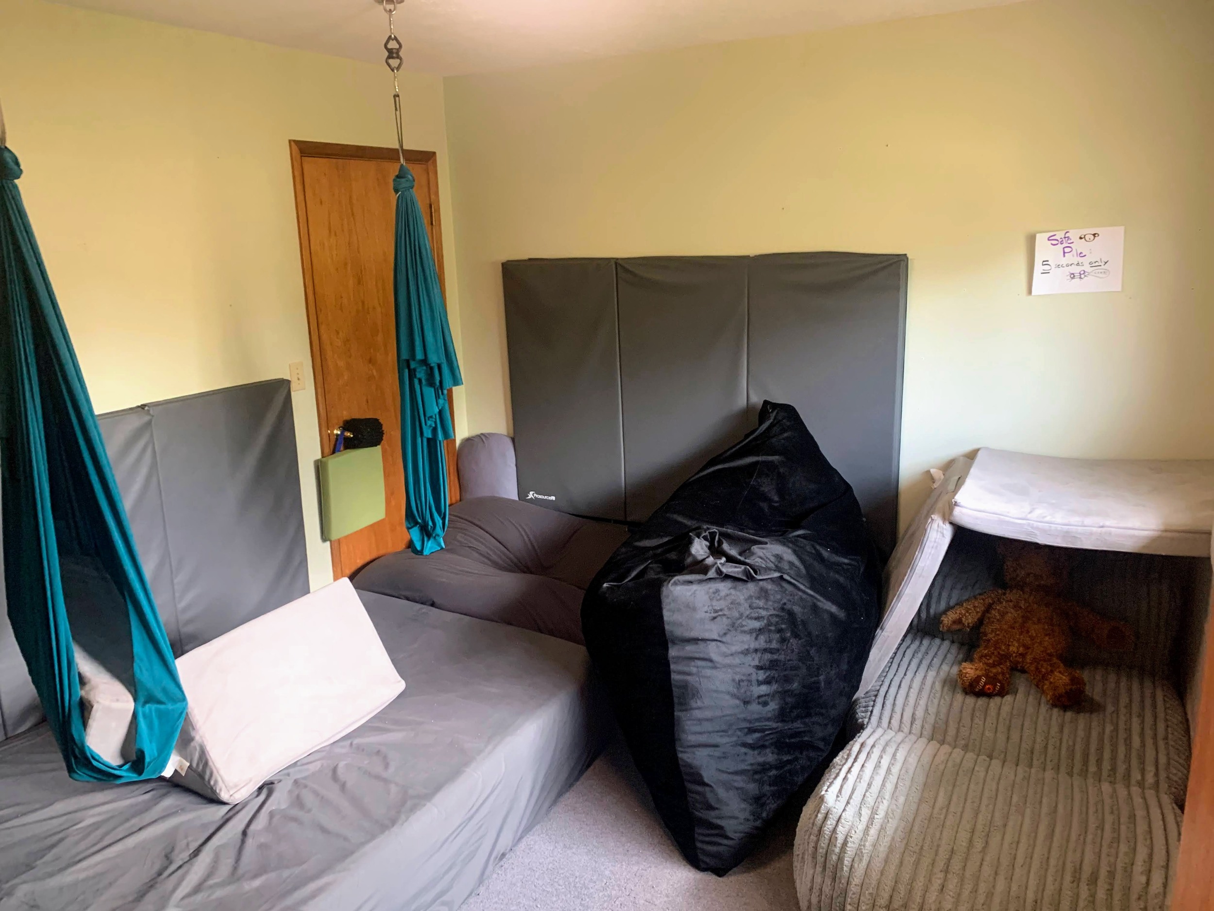 A bedroom with two beds, one with a white pillow and the other with a brown teddy bear, black clothing, a black speaker, and blue aerial yoga hammocks hanging from the ceiling. A cardboard sign on the wall reads 'Safe Place' with handwritten notes and drawings.
