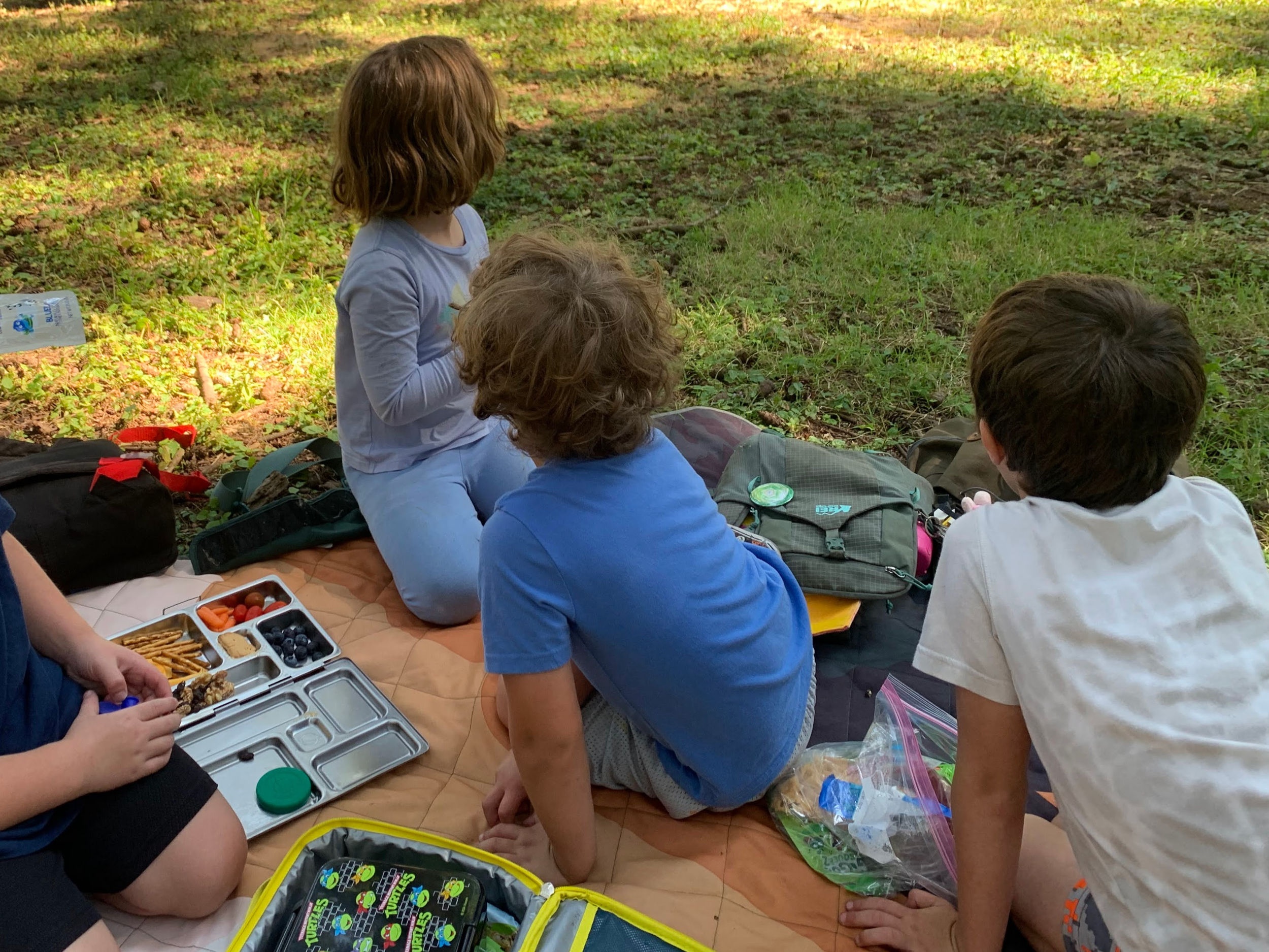 Children sitting on a blanket outdoors during lunch, surrounded by backpacks and lunch containers, with trees and grass in the background.