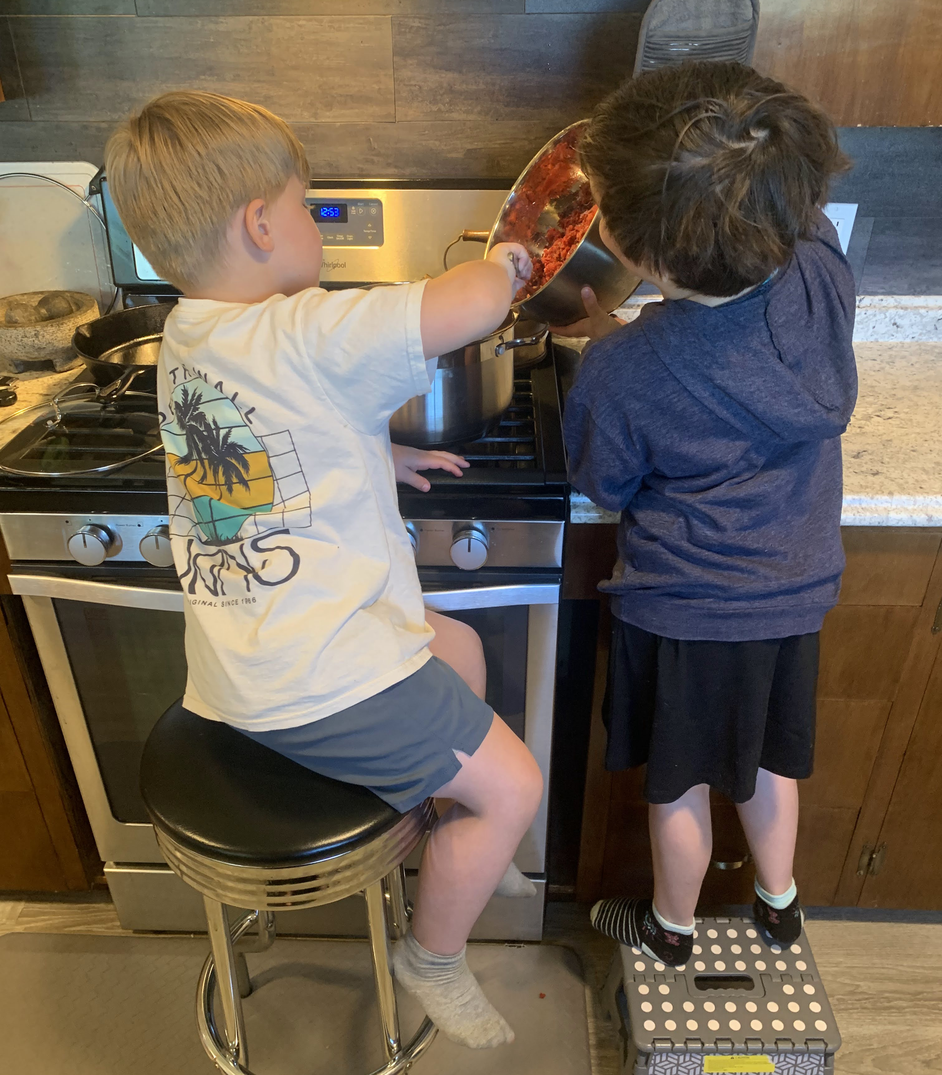 Two young boys standing on a step stool, measuring homemade tomato sauce and pouring it into a large pot on a kitchen stove in a home kitchen with wooden cabinets and tiled walls.