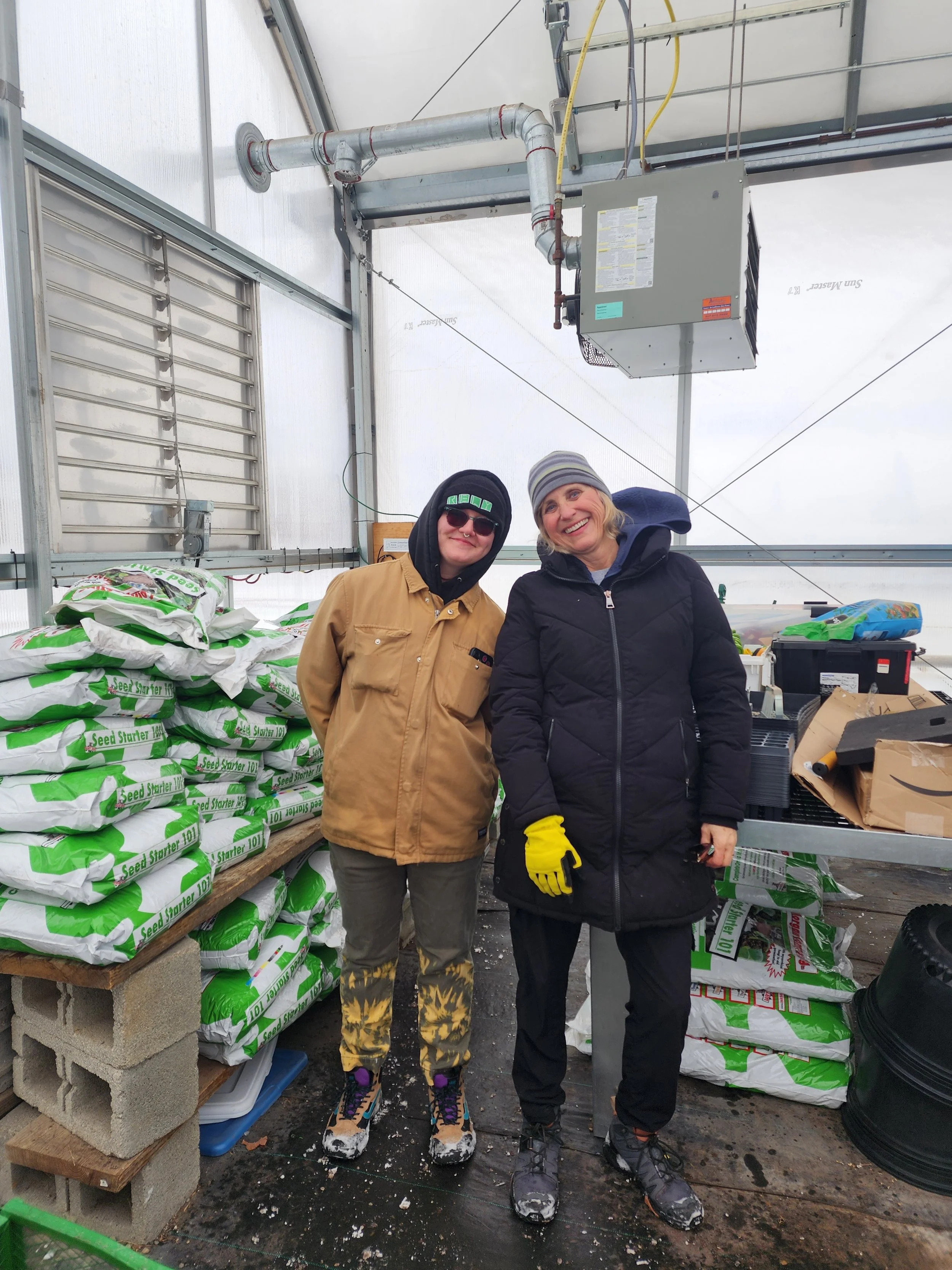 Two people in jackets, smiling, with compost bags in a greenhouse