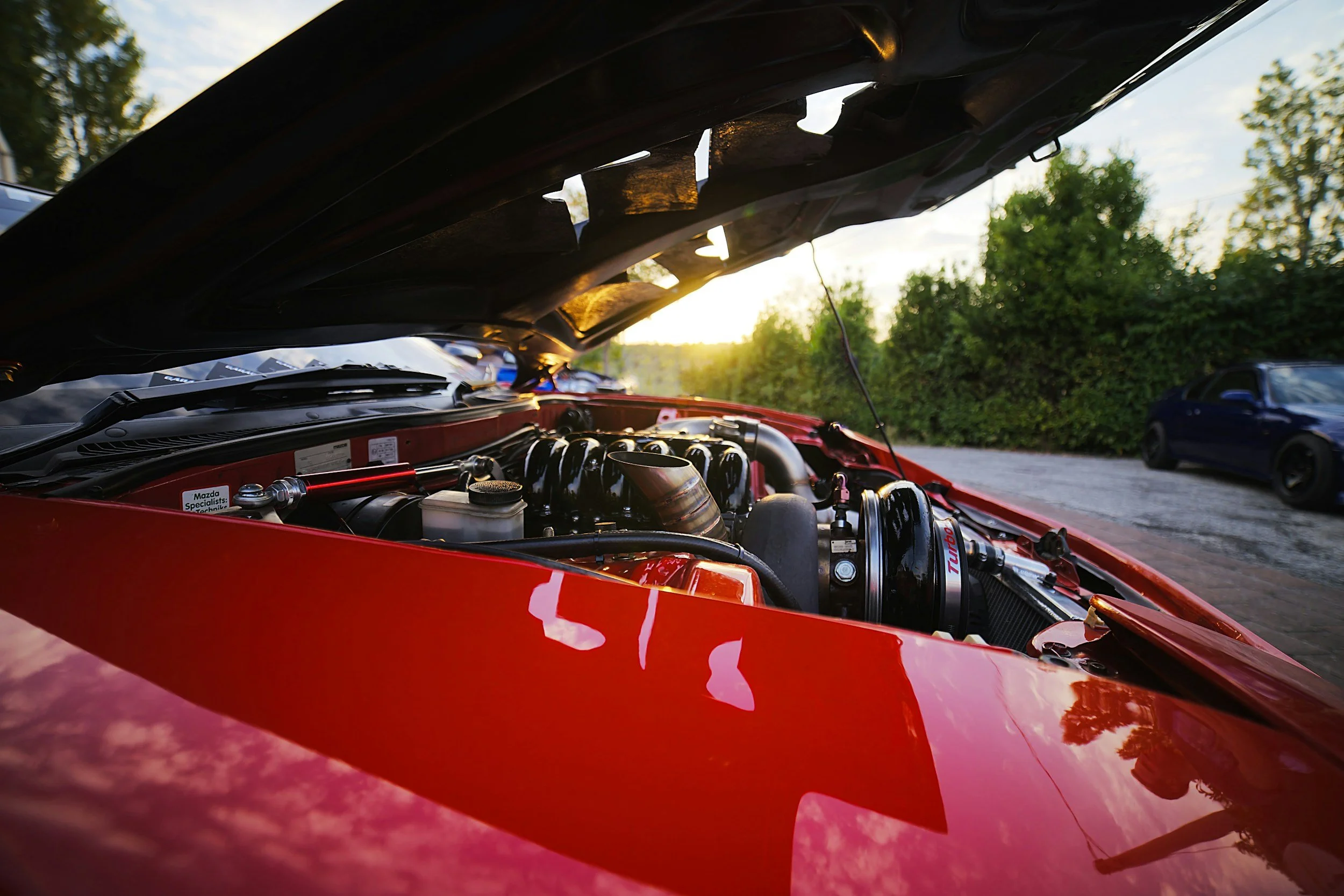 Closer view of a red sports car with open hood revealing engine components, parked outdoors during sunset with greenery and a blue car in the background.
