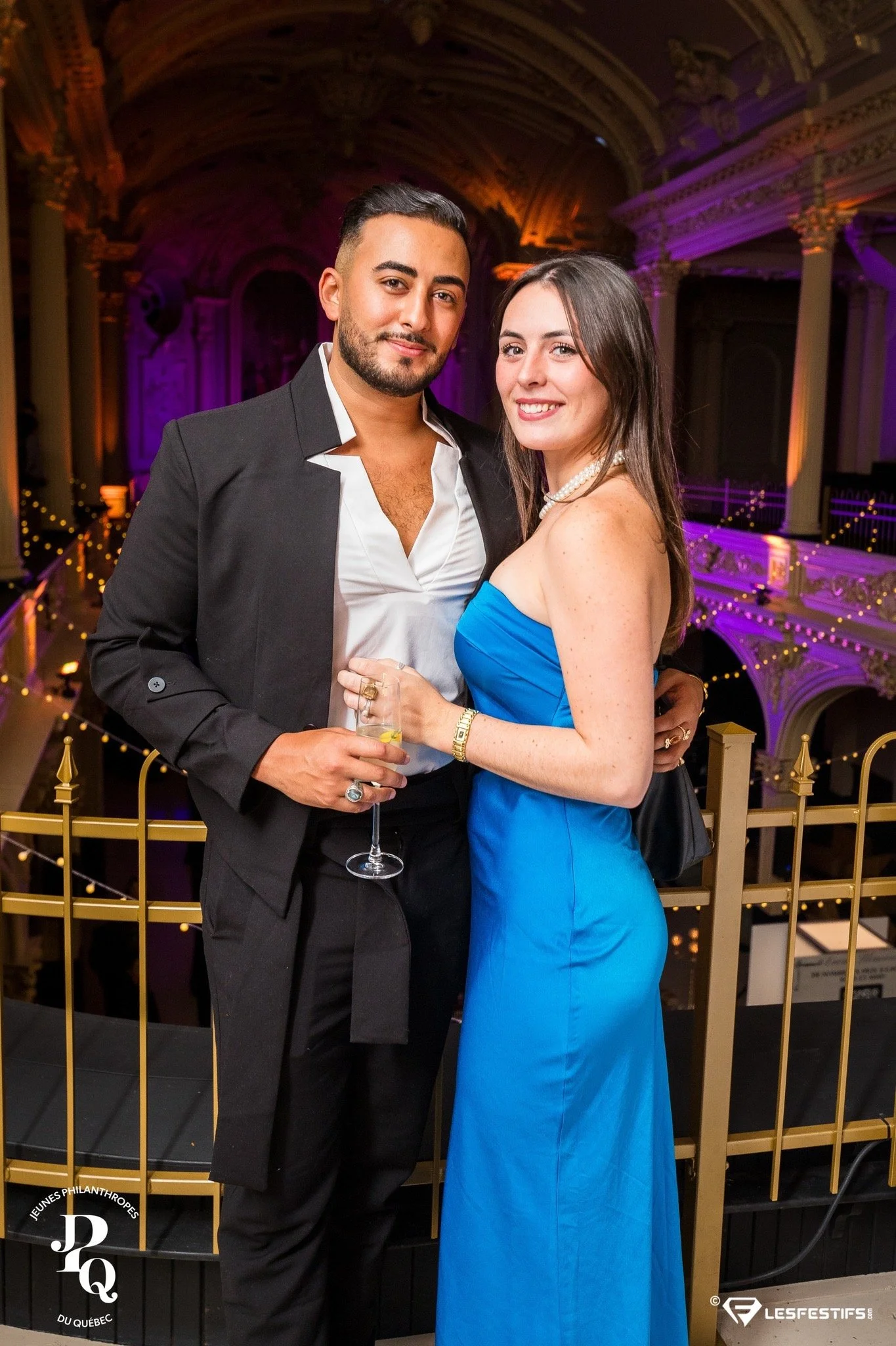 Un couple bien habillé posant pour une photo dans un lieu élégant avec des lumières violettes. L'homme porte un costume noir et une chemise blanche, la femme porte une robe bleue. Ils tiennent un verre à la main.