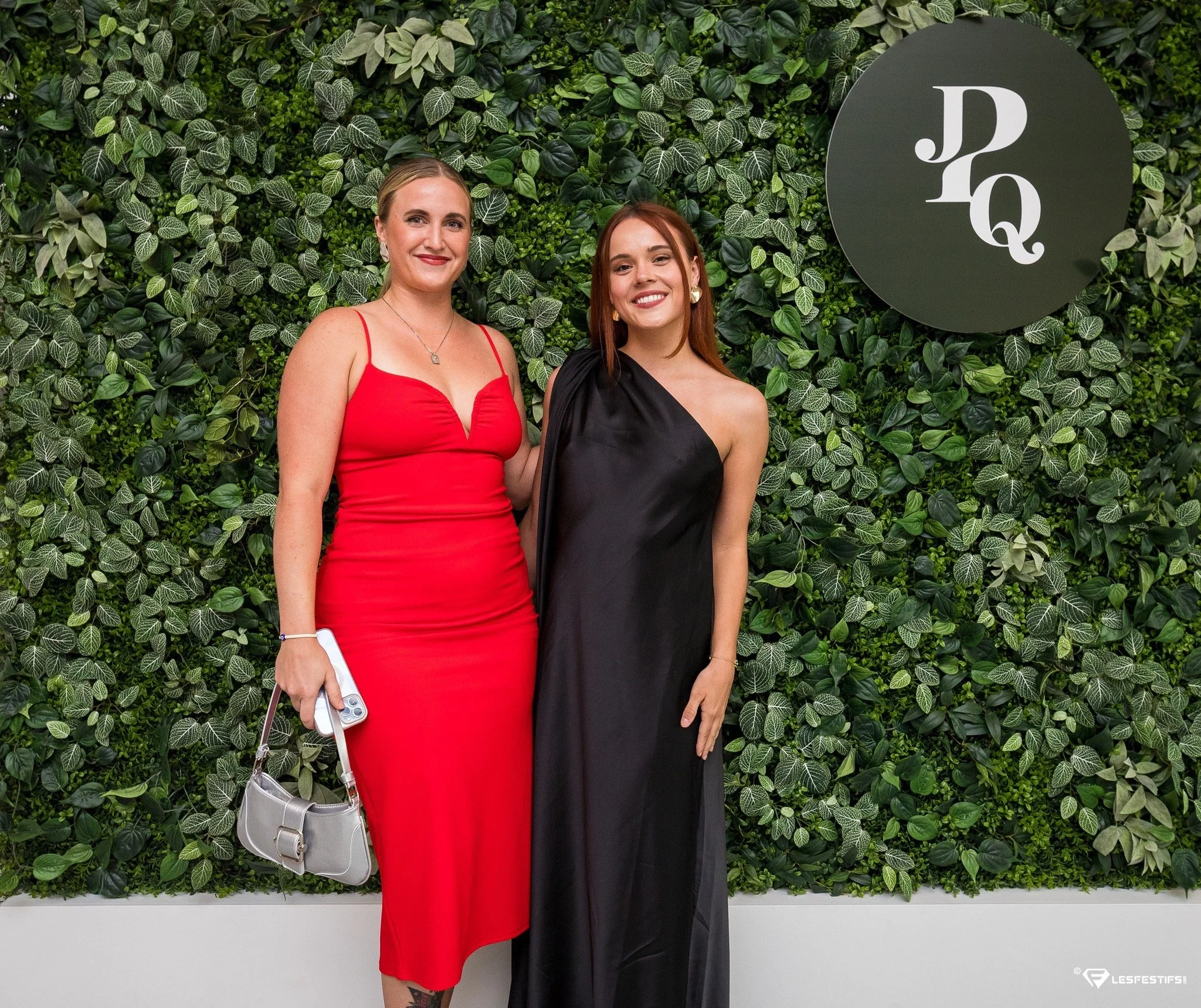 Deux femmes posent devant un mur de feuilles vertes avec le logo 'JQ'. L'une porte une robe rouge et l'autre une robe noire. Elles sourient.