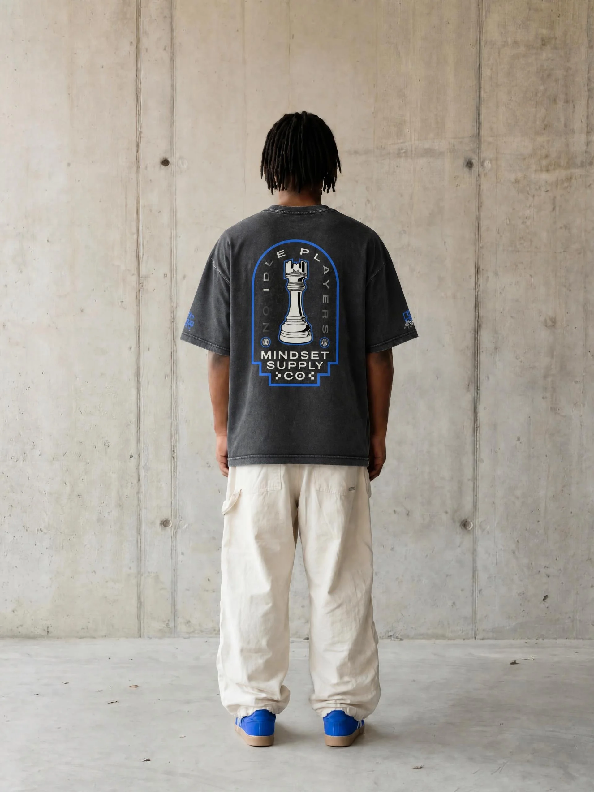 Person with dreadlocks facing a plain concrete wall, wearing a black T-shirt with a chess piece logo and text, paired with white cargo pants and blue sneakers.