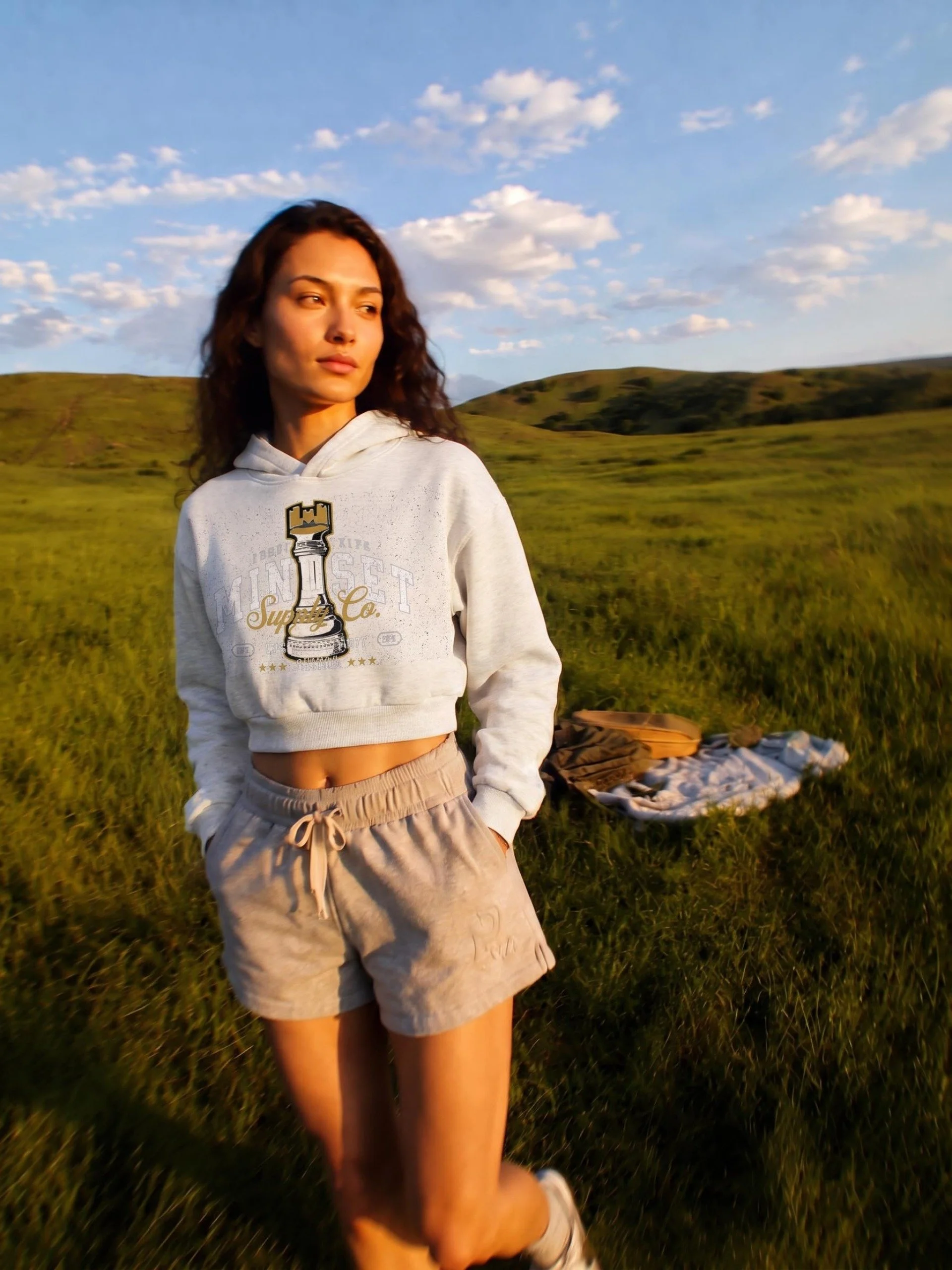 A young woman with curly brown hair and tan skin standing in a grassy field under a blue sky with scattered clouds, wearing a white hoodie with a chess piece graphic and the words 'Super Co.' and beige shorts.