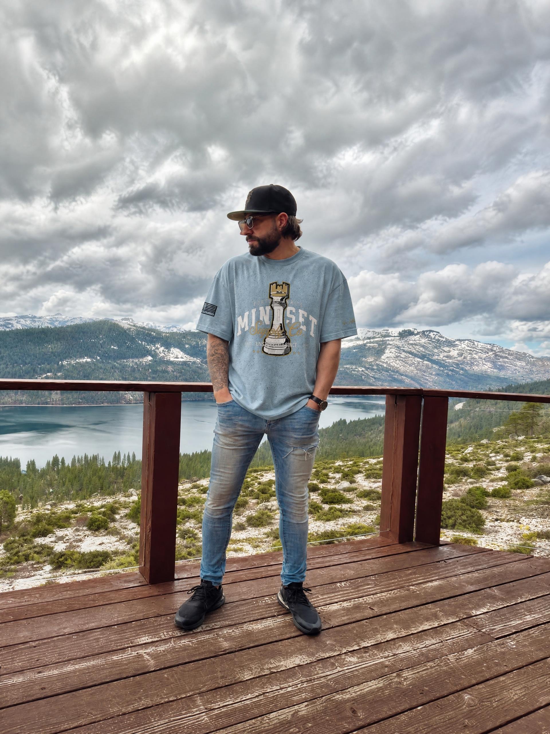 A man with sunglasses and a black cap standing on a wooden deck overlooking a mountain landscape with cloudy skies, water, and snow-capped peaks in the background.