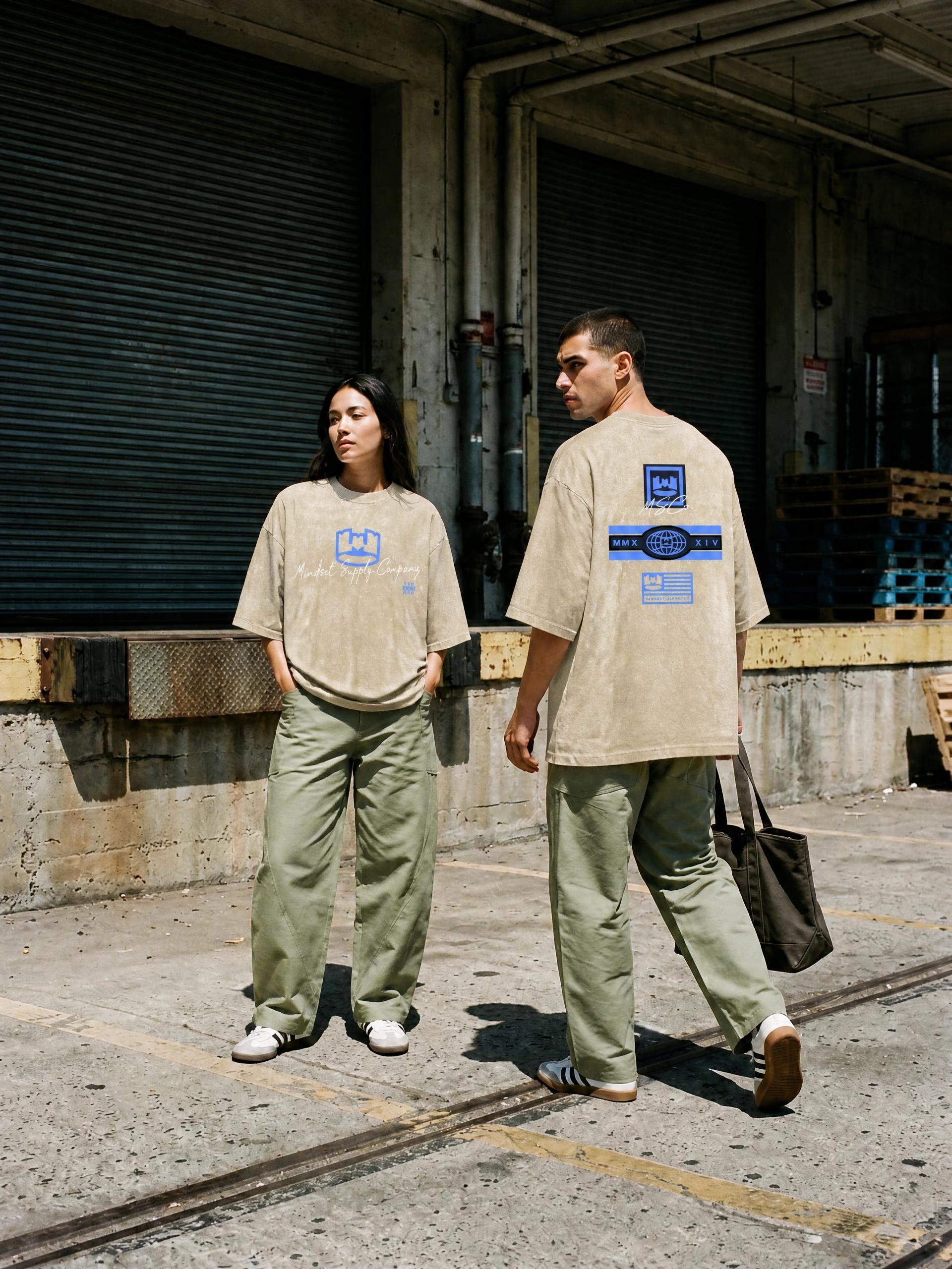 Two young adults, a woman and a man, standing outdoors against a concrete wall with a black metal shutter, wearing matching beige oversized T-shirts and baggy green pants, with the woman having her hands in her pockets and the man holding a black bag
