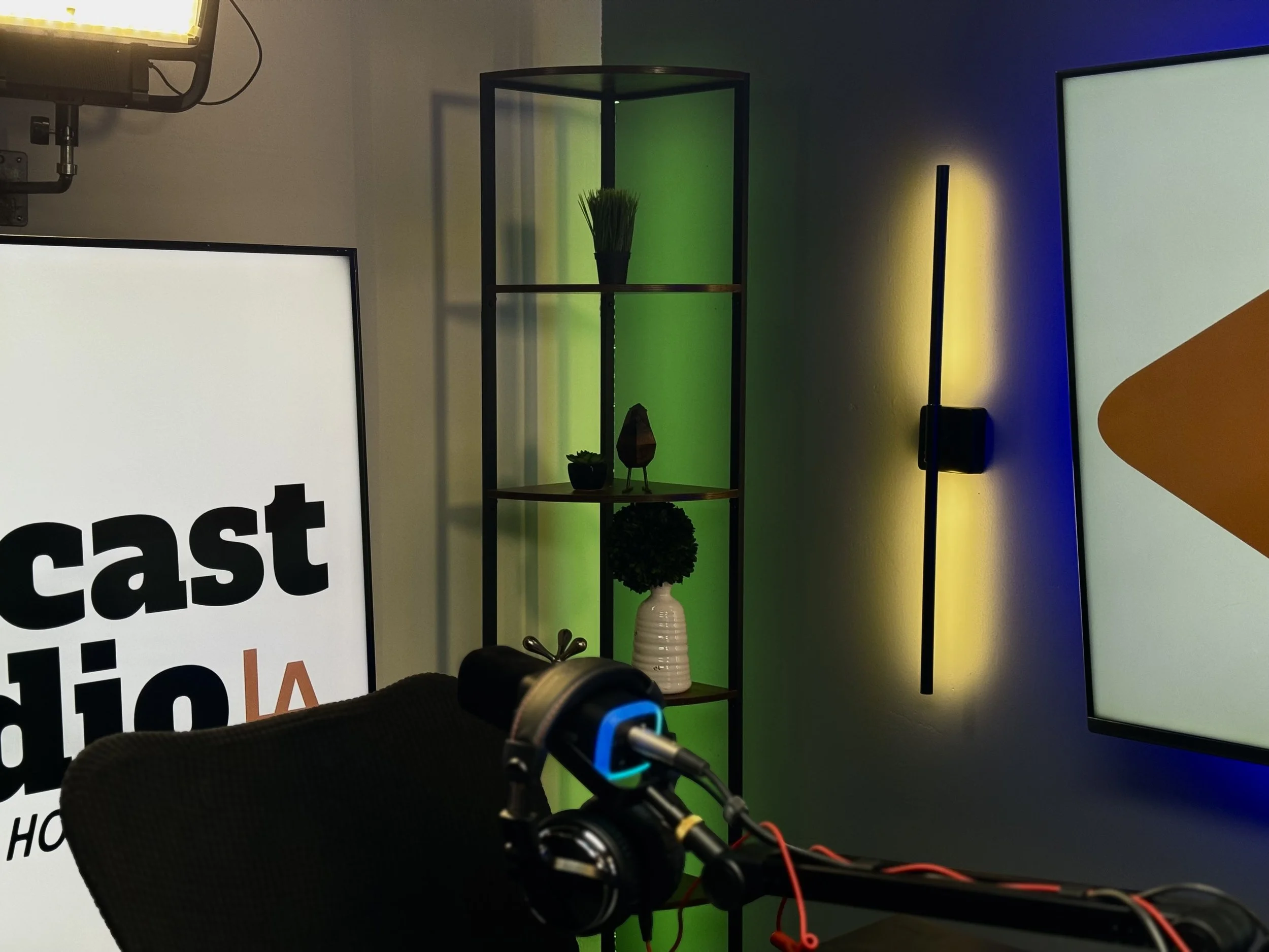 A television news studio with a monitor displaying the word "cast" and the partial word "radio", a microphone, a black chair, a modern black ladder shelf with decorative items, and colorful LED lighting on the wall.