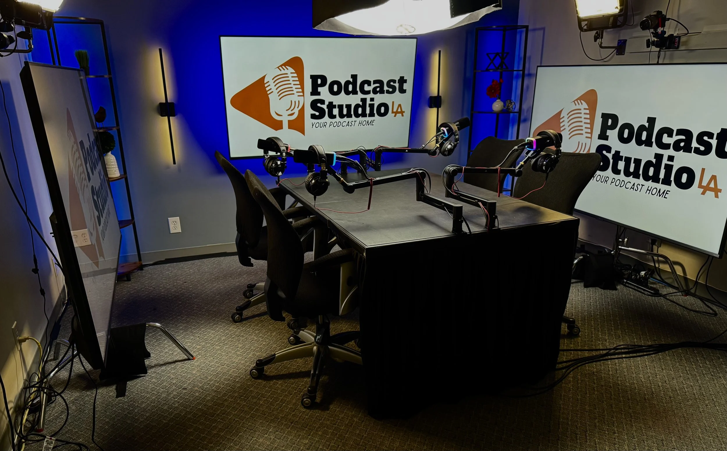 A podcast recording studio with a table and chairs, two large screens displaying the Podcast Studio LA logo, microphones with headphones, and professional lighting.