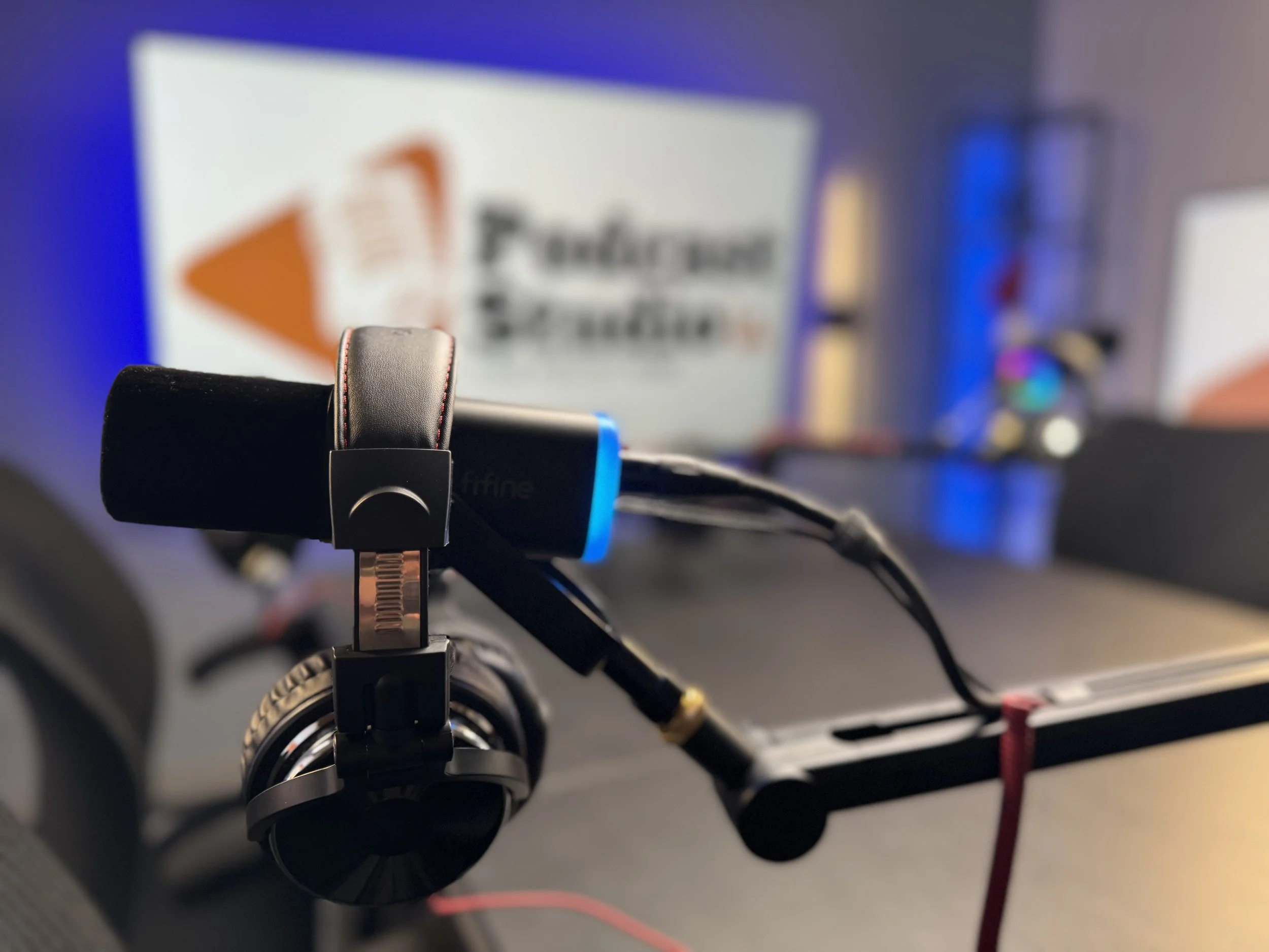 Close-up of headphones and a microphone on a desk with blurred monitors in the background.