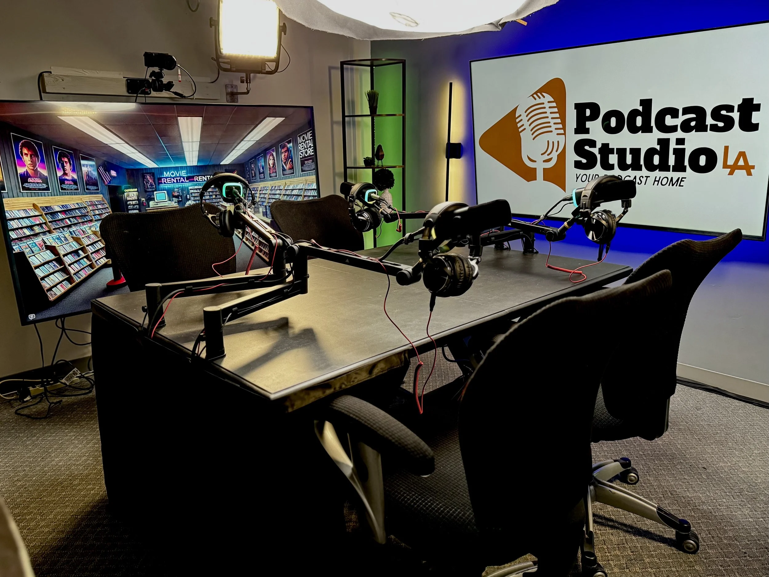Podcast studio with a large screen displaying 'Podcast Studio LA' with a microphone logo, a table with four microphones and headphones, and a second monitor showing a bookstore scene with movie posters.