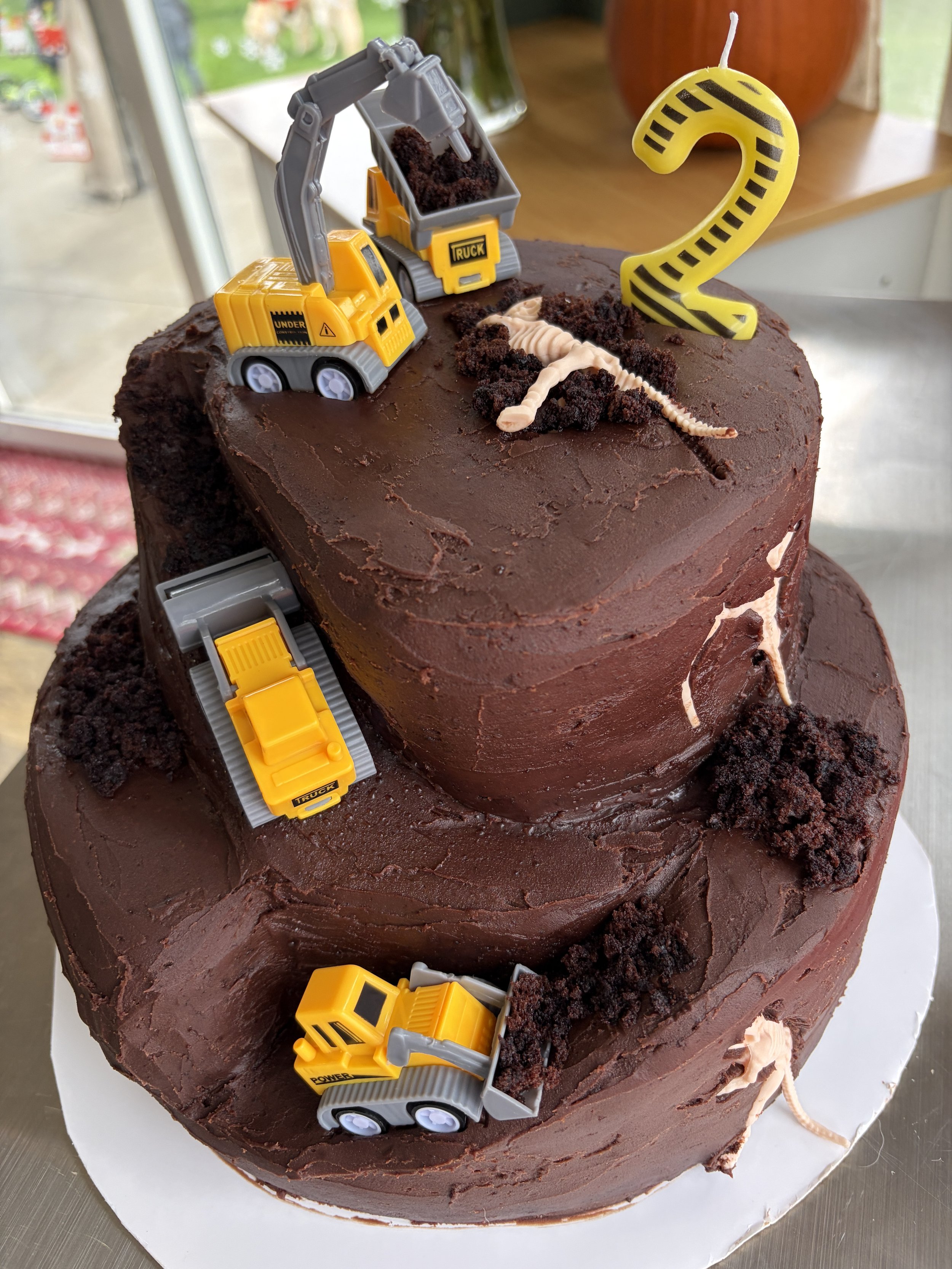 Chocolate construction cake topped with plastic dinosaurs, construction vehicles, and a number two candle.