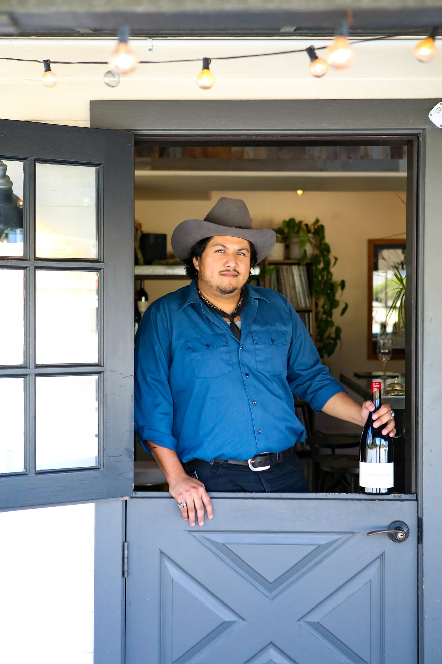Vince with a cowboy hat, proudly holding a bottle above the barn style door to the tasting room.