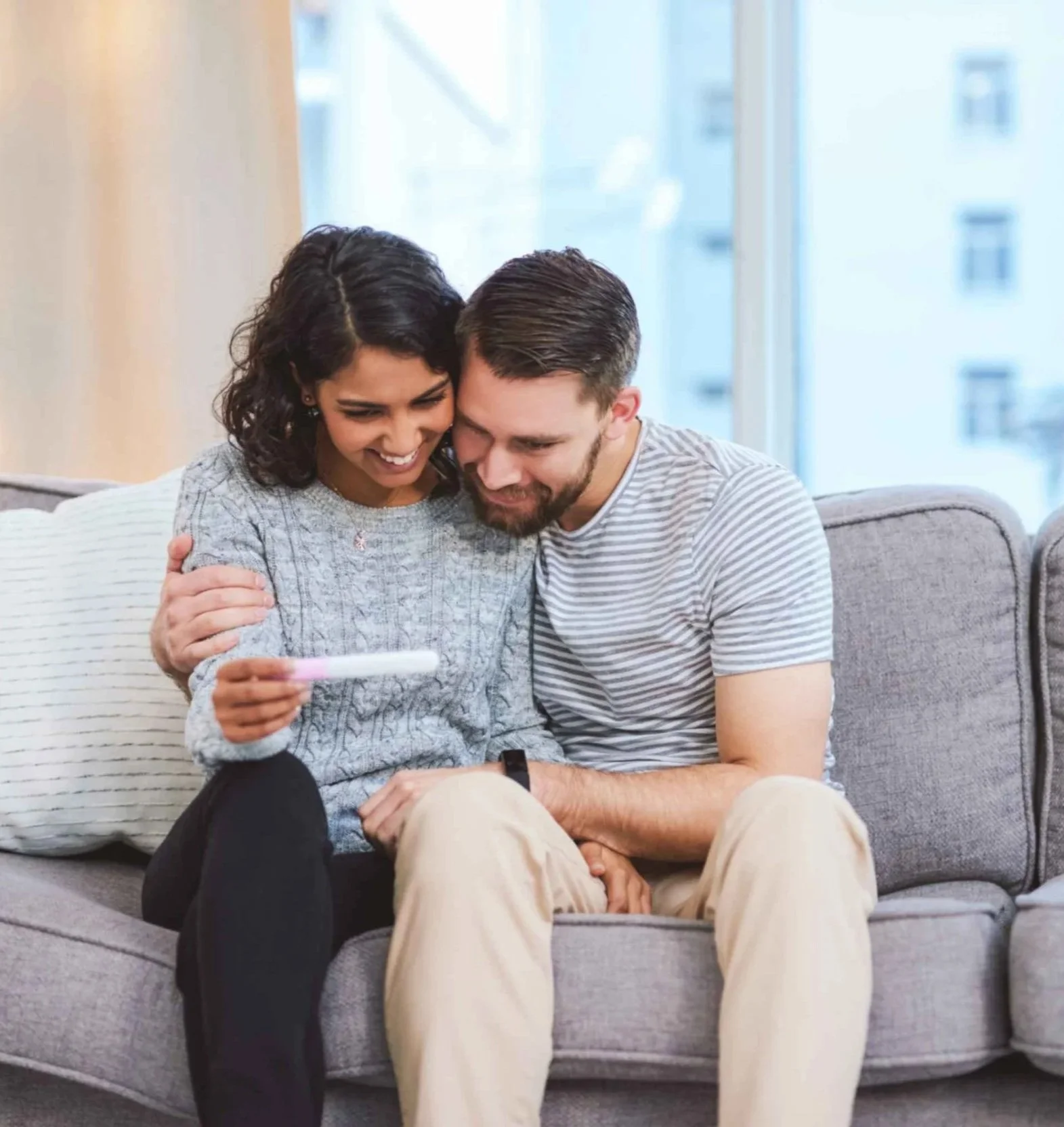 Young female holds pregnancy test and smiles while sitting on couch with partner who is hugging her
