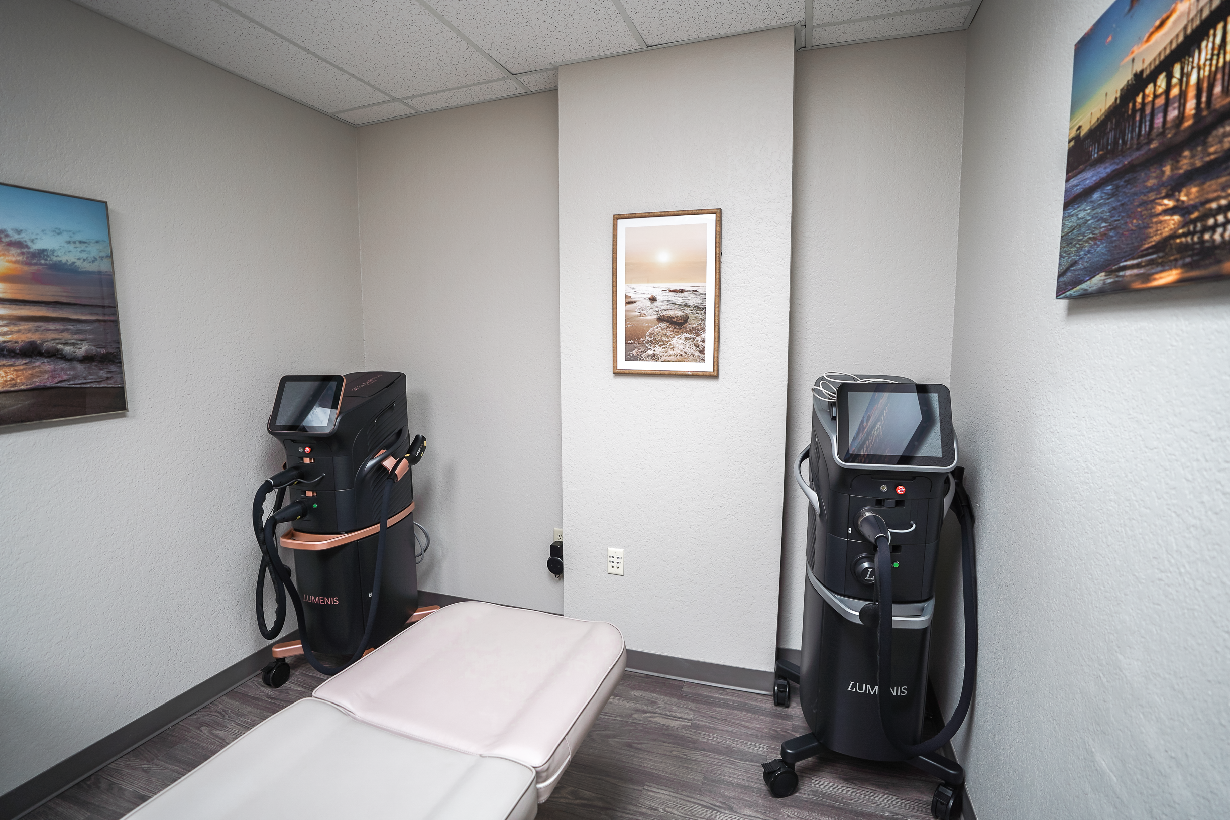 A small examination room with two medical devices and a padded examination table. One wall has a framed picture of a beach scene, and two wall-mounted screens display sunset beach images. The room has gray walls, a gray floor, and a white ceiling with ceiling tiles.
