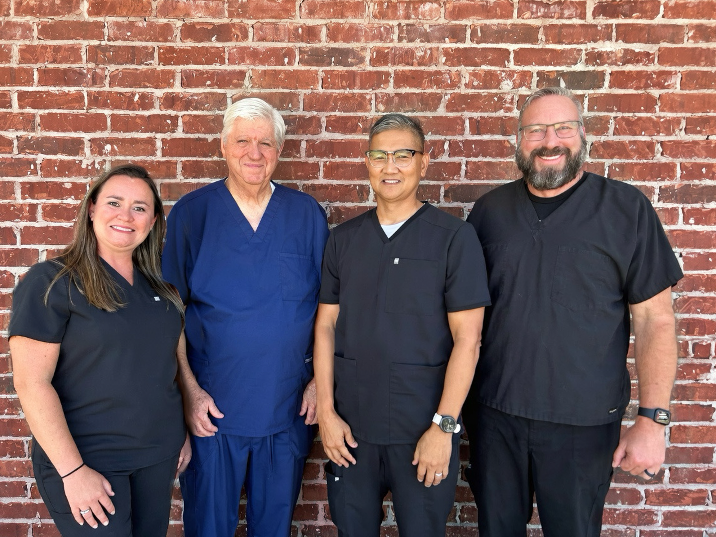 Group of five healthcare professionals standing in front of a red brick wall, wearing medical scrubs and smiling.