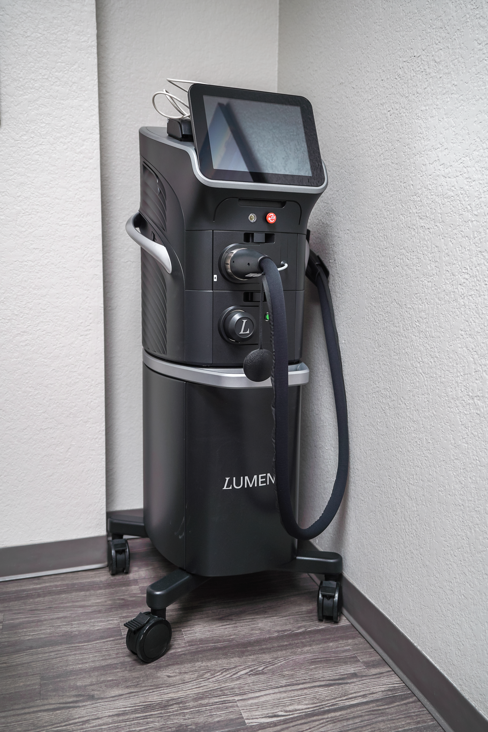 A medical laser machine labeled 'LUMEN' positioned in a corner of a room with light-colored walls and dark wooden flooring.