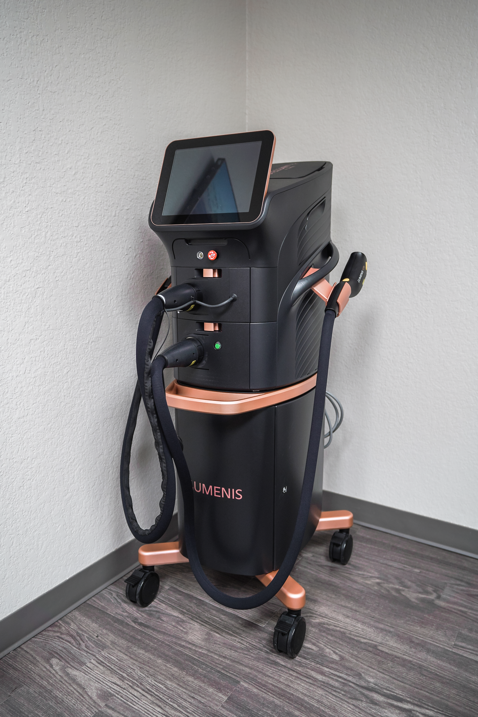 Medical device on a wheeled stand with a touchscreen display, black hoses, and a black and pink color scheme, positioned against a beige wall in a clinical setting.