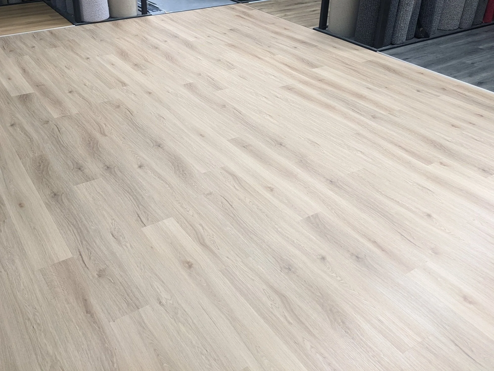 Wood grain laminate flooring in a store setting with carpet rolls in the background.
