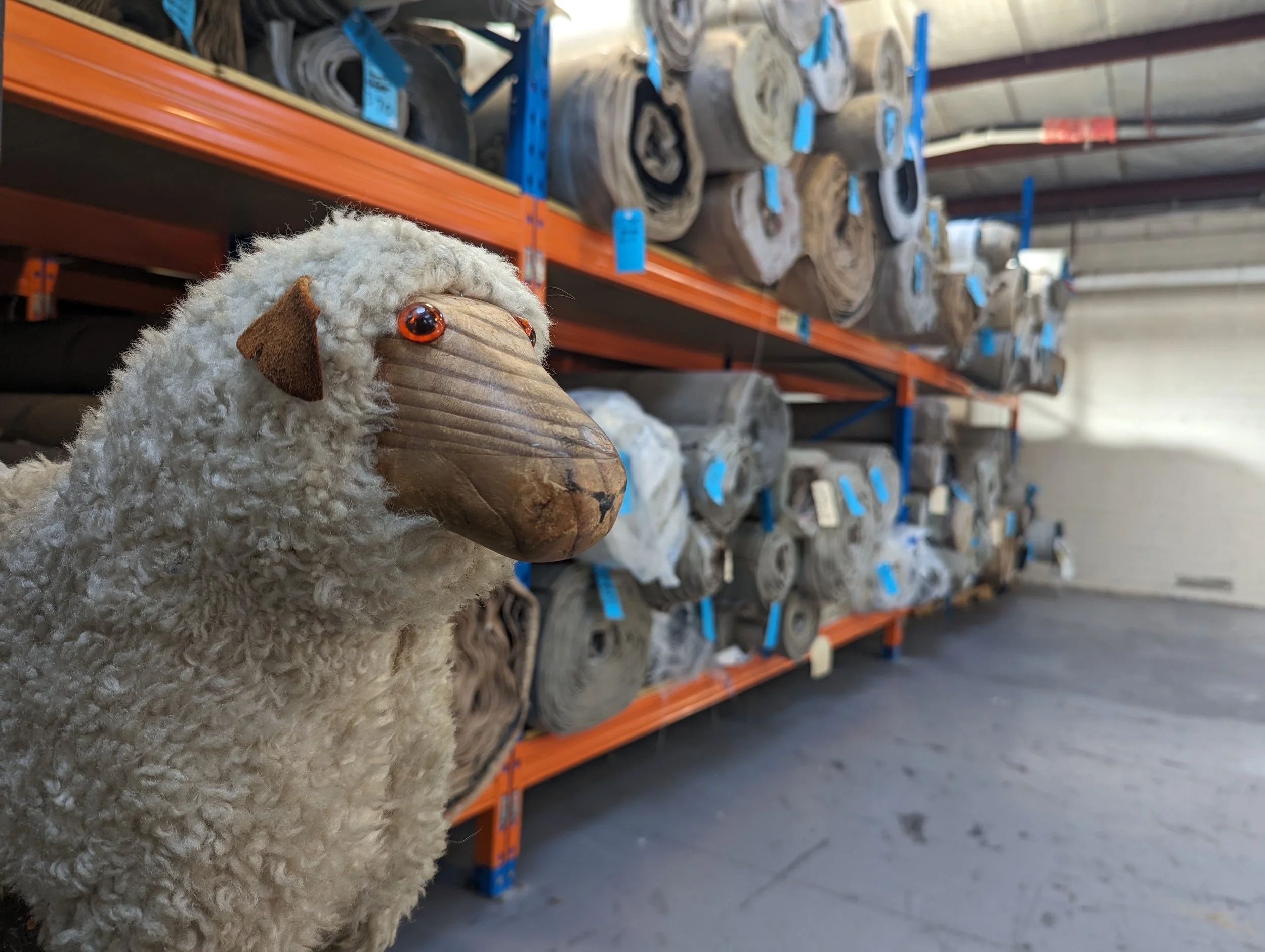 Stuffed toy sheep in a warehouse with rolls of carpet on racks.