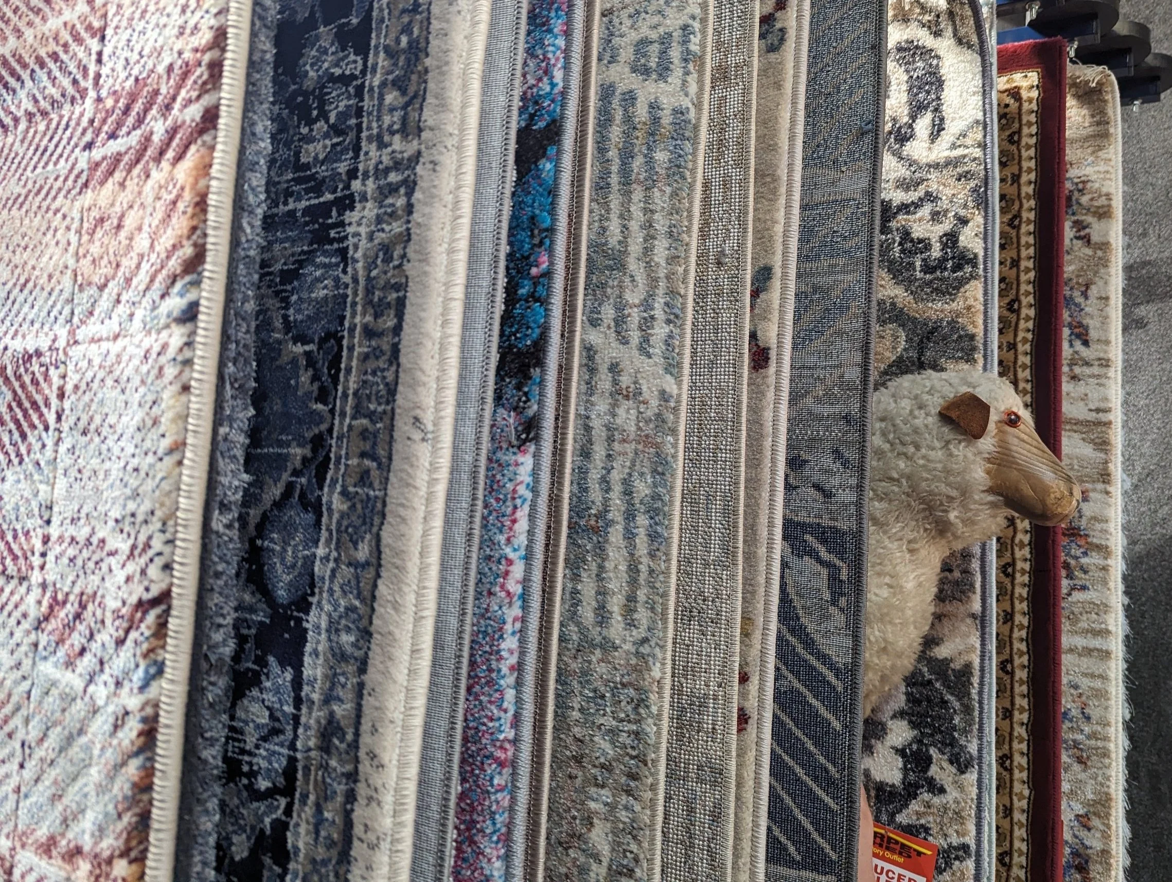 Various patterned rugs hang vertically in a display, with a stuffed toy sheep partially visible among the rugs.