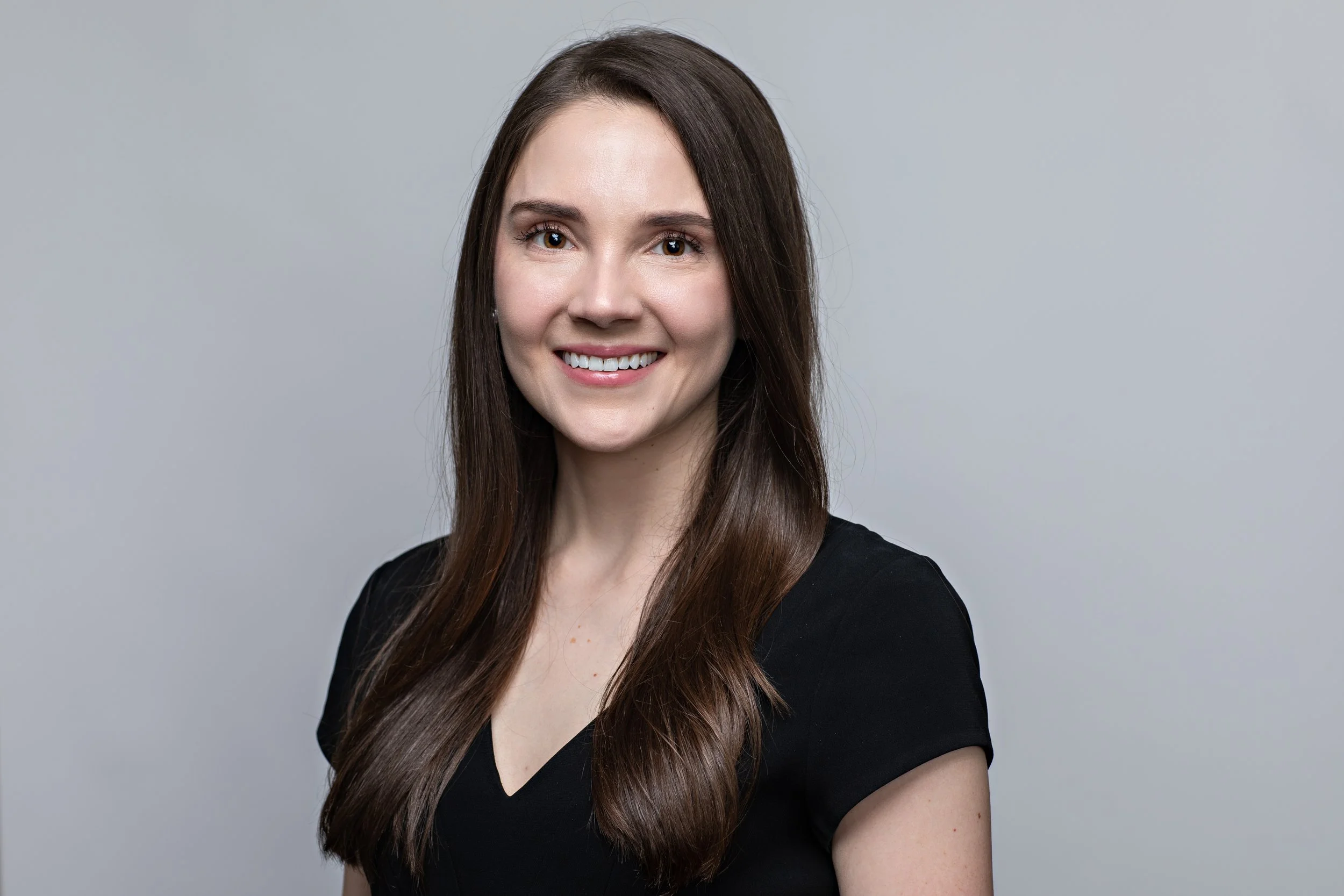 A young woman with long brown hair wearing a black top, smiling with white teeth, against a plain gray background.