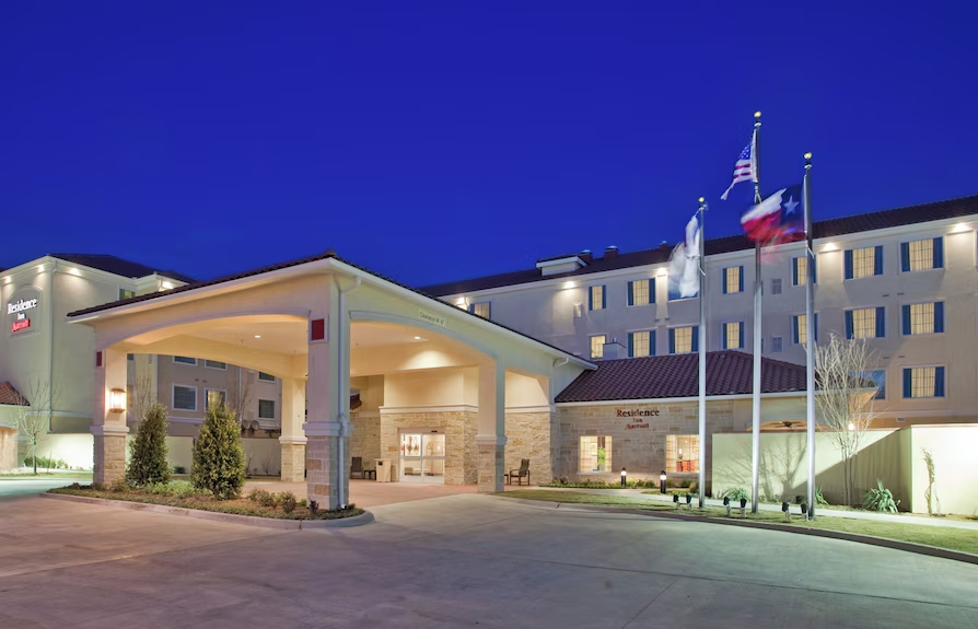 Exterior view of a hotel at night with illuminated building, flagpoles with American, Texas, and other flags, and a covered entrance.