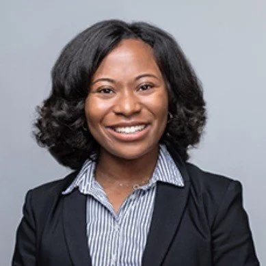 A professional woman with shoulder-length black hair, wearing a striped button-up shirt and a black blazer, smiling at the camera against a plain gray background.